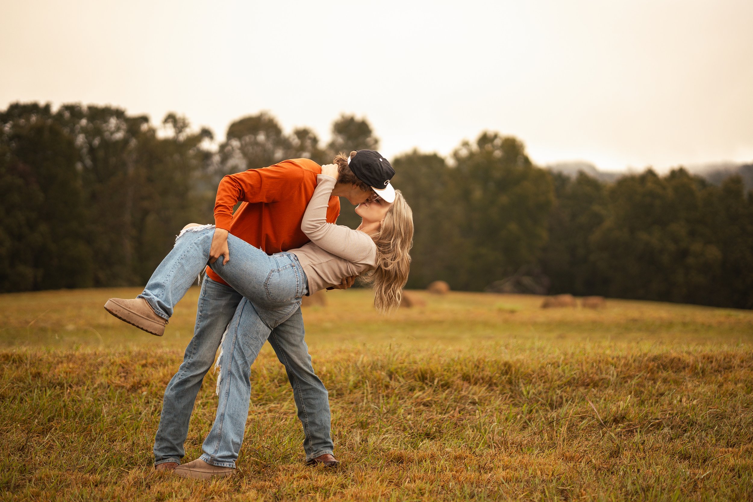 A man is carrying a woman in his arms while kissing outdoors in a grassy field with trees in the background.