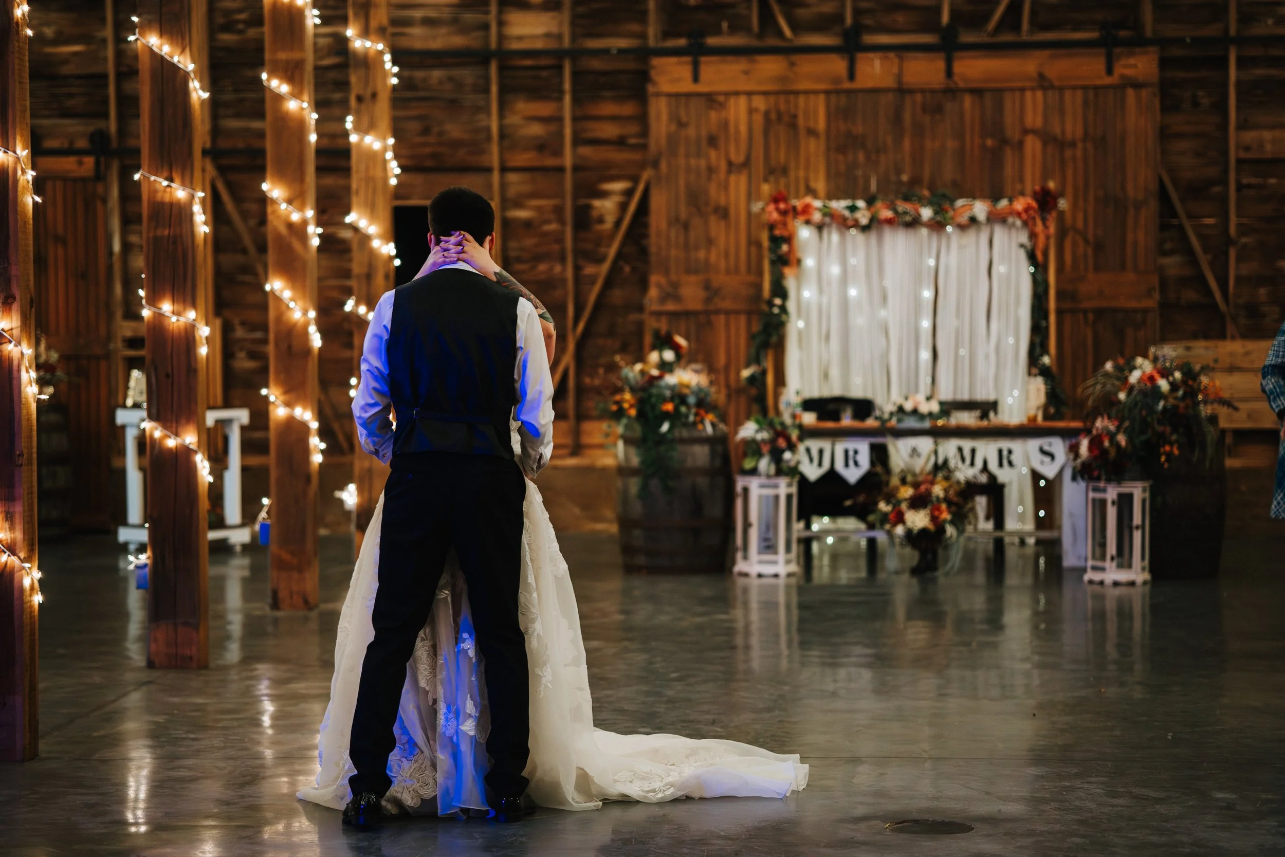 A couple sharing a dance at their wedding reception in a rustic barn decorated with string lights and floral arrangements.