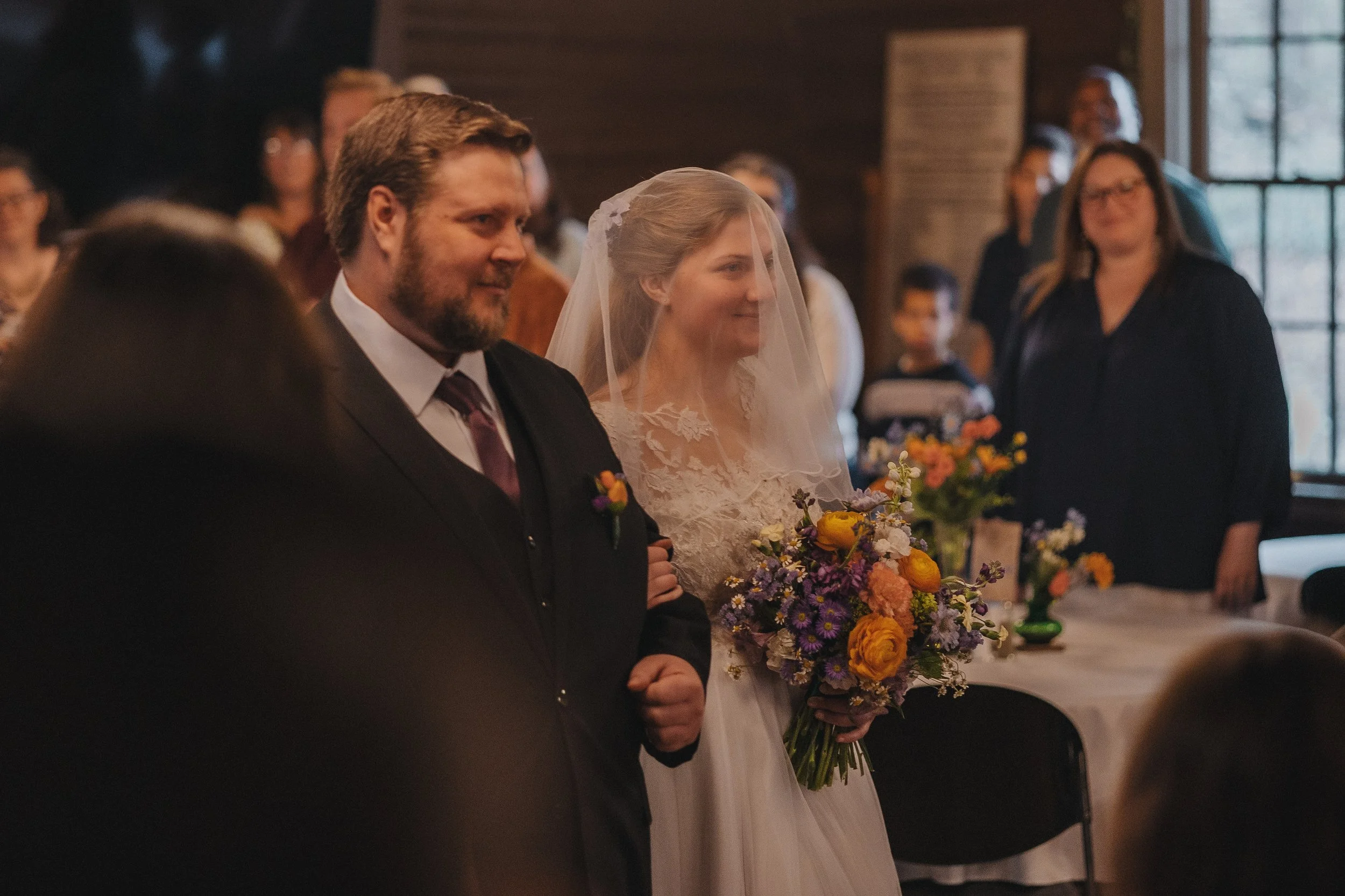 A bride and groom standing together during a wedding ceremony, with guests in the background, indoors with wooden walls and large windows, the bride holding a colorful bouquet of flowers.