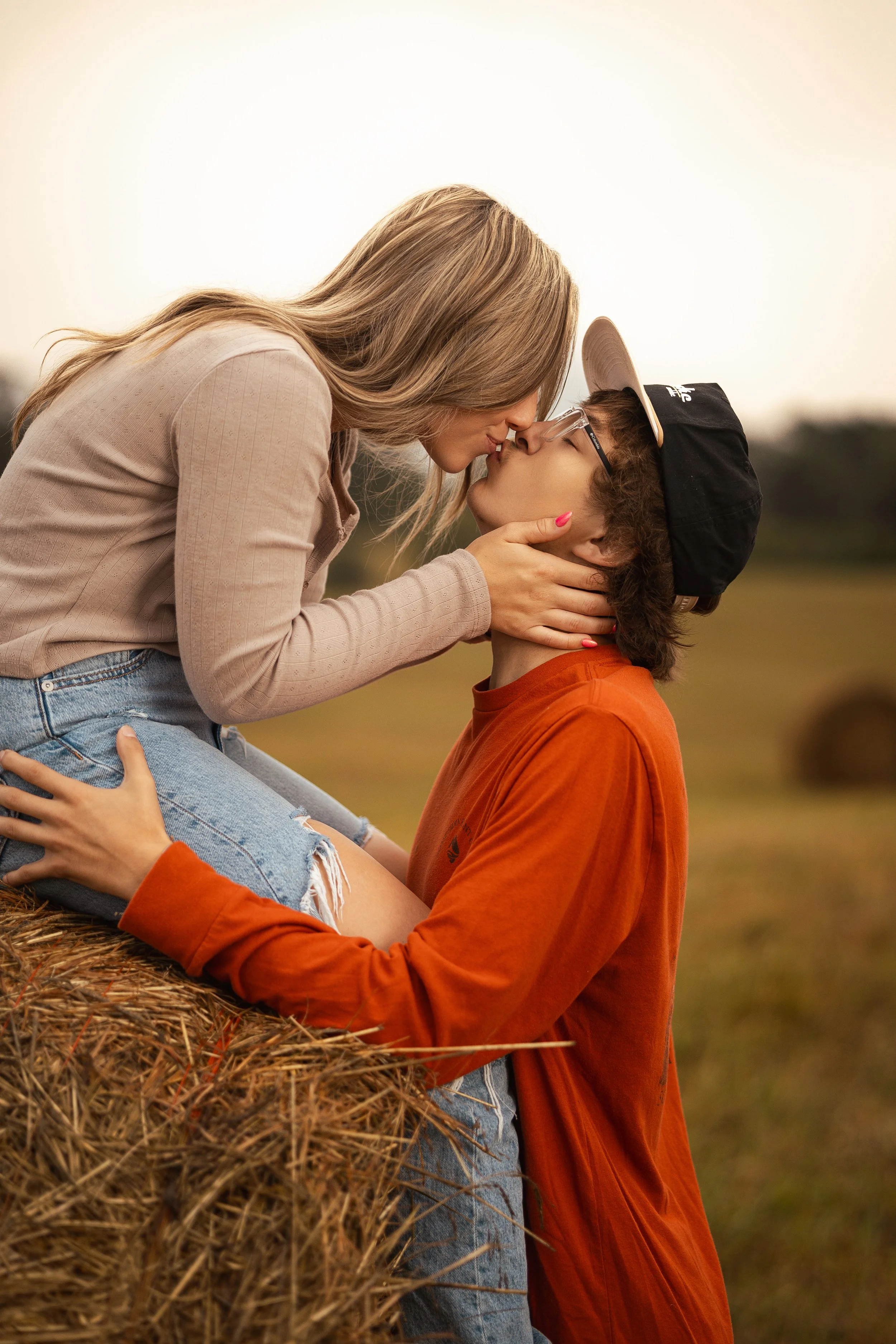 A woman and a young man kiss each other outdoors near a hay bale during sunset.