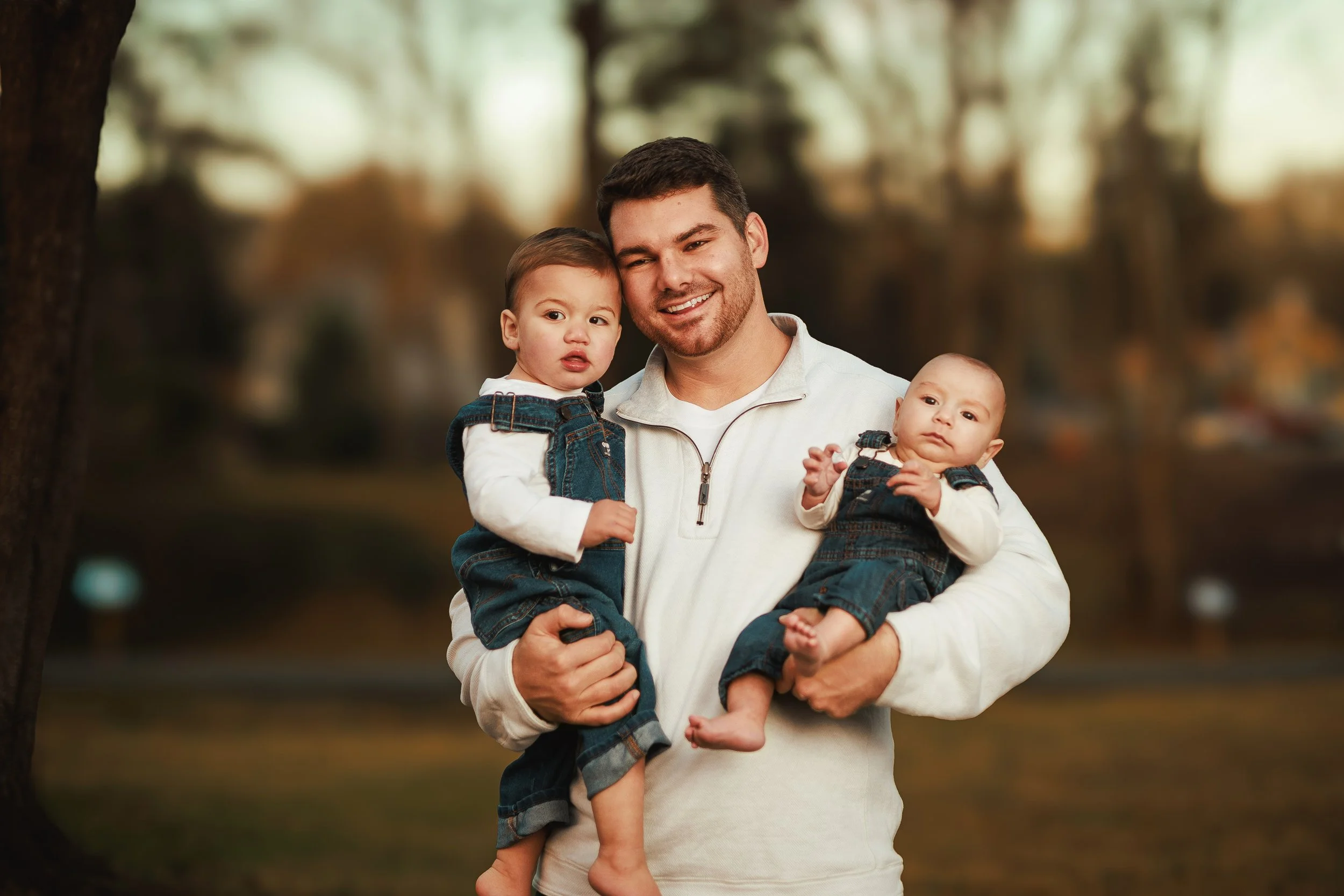 A happy man holding two children, one in each arm, outdoors in a park with blurred trees in the background during autumn.