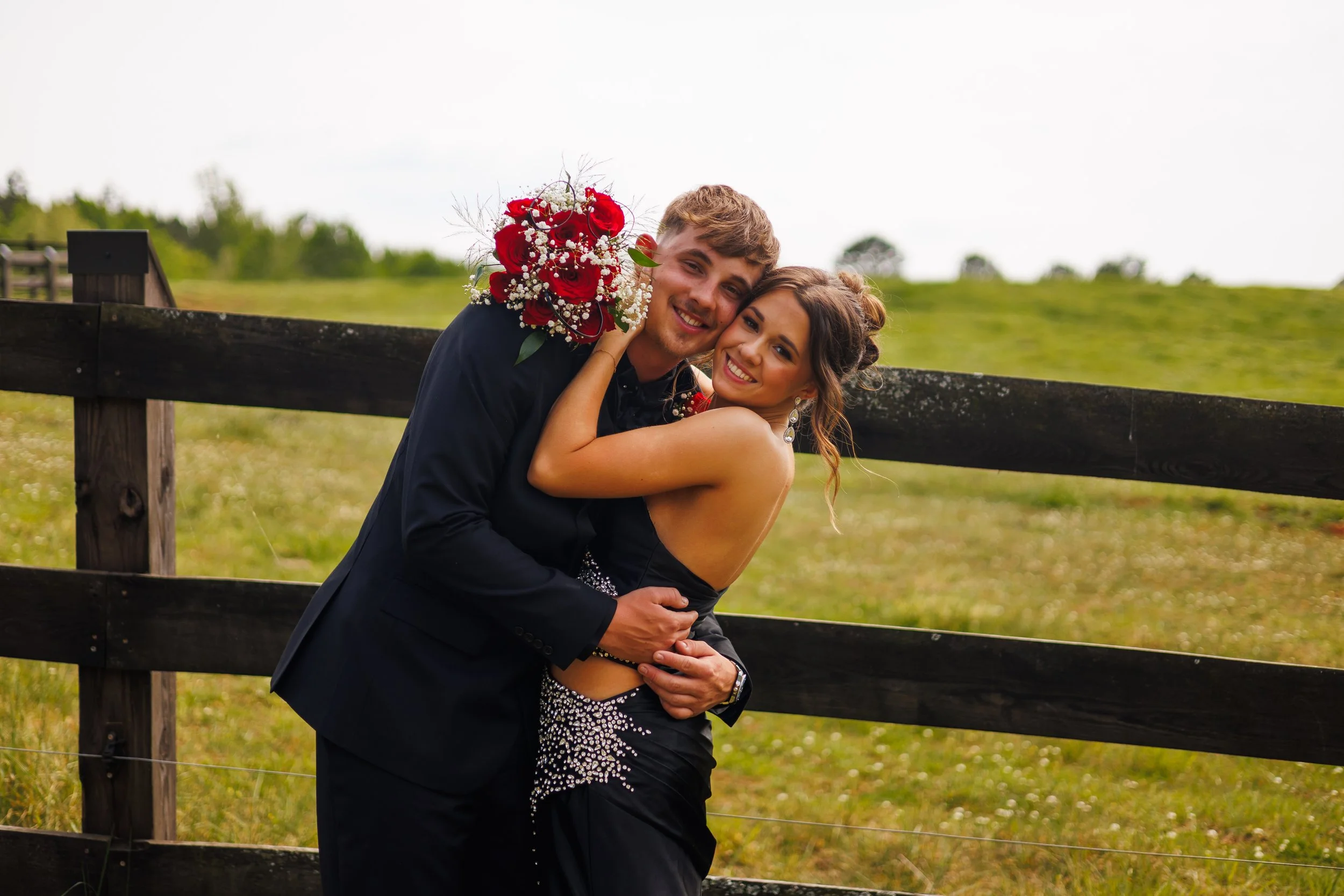 A happy couple embracing outdoors, the man holding a bouquet of red and white flowers, standing in front of a wooden fence with a grassy field and trees in the background.
