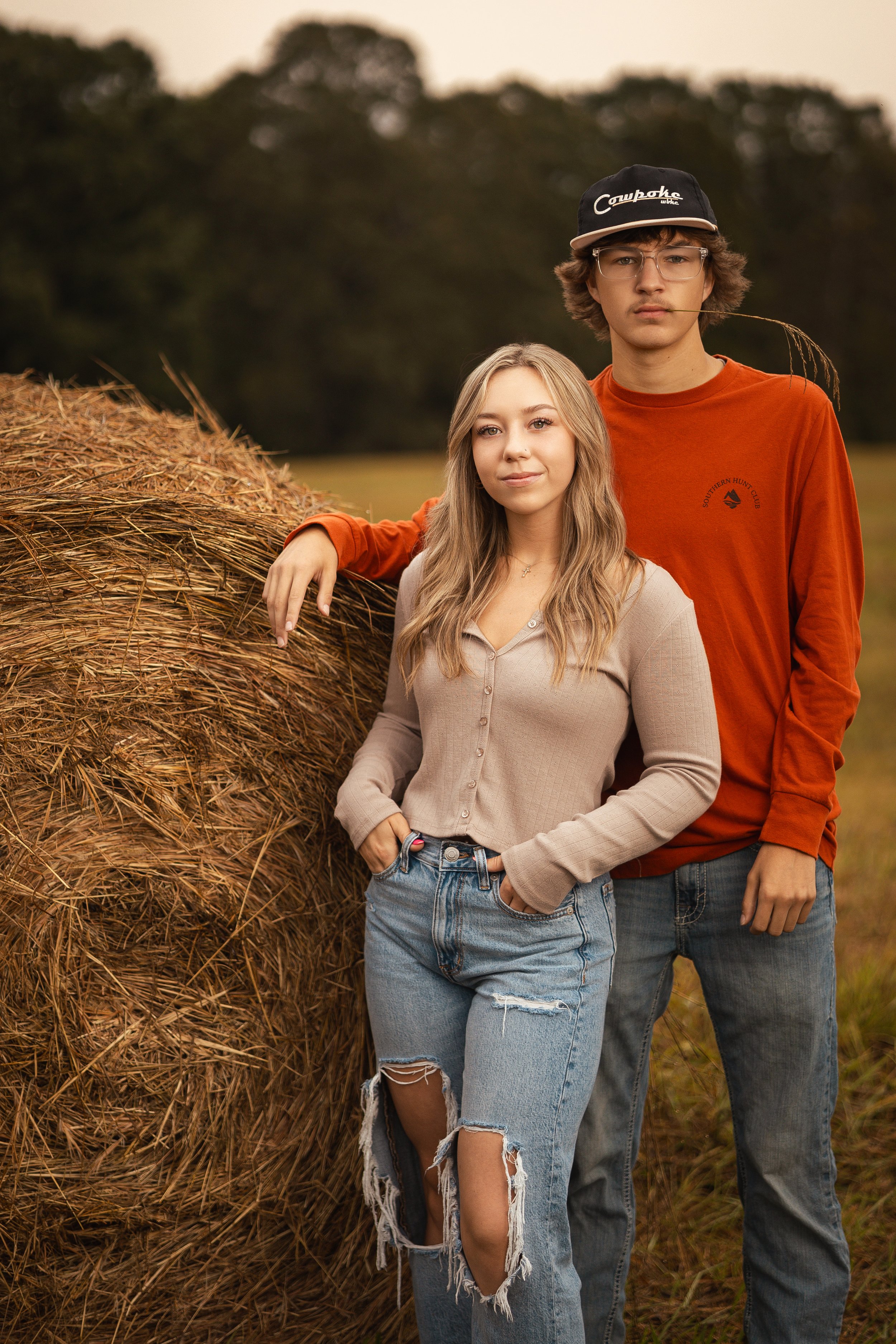 A young woman and a young man standing outdoors next to a large hay bale, with trees in the background, during what appears to be early evening or sunset.