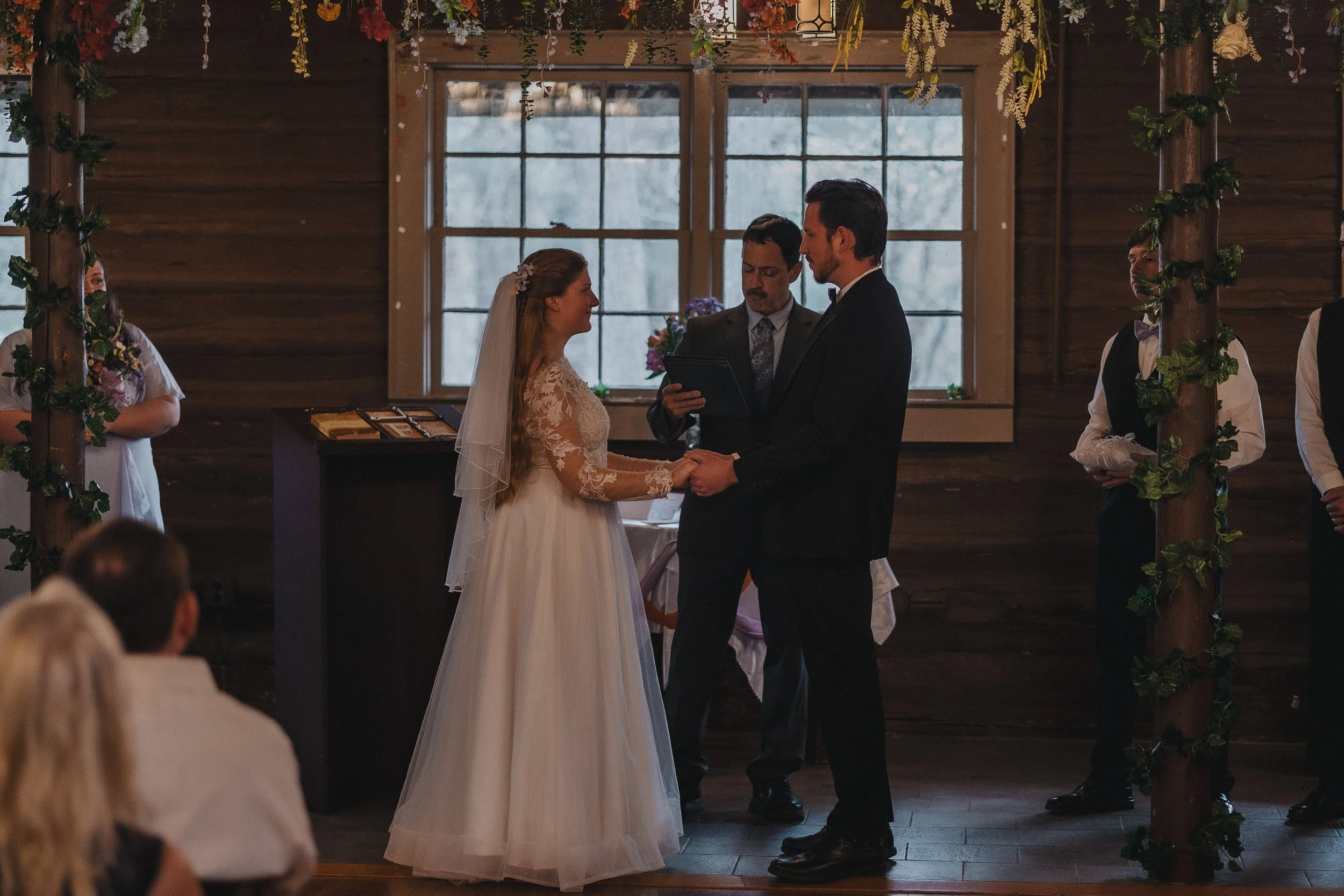 A bride and groom holding hands and exchanging vows during a wedding ceremony in a rustic wooden venue with large windows and floral decorations.