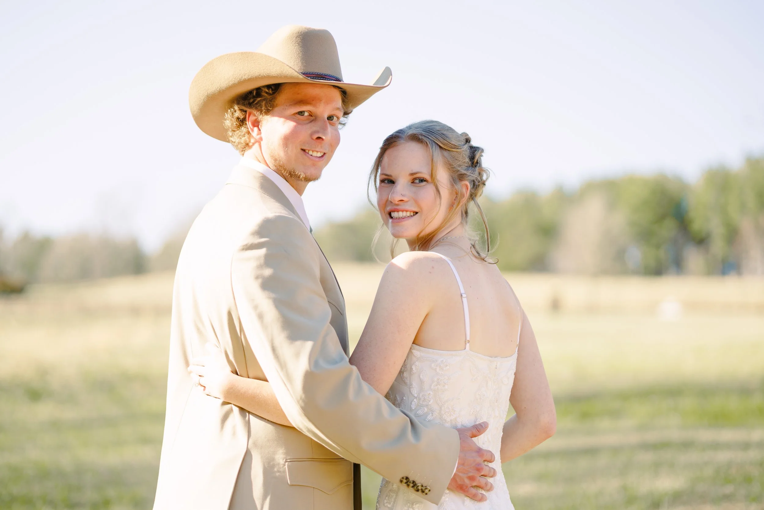A couple in wedding attire standing outdoors, with the man wearing a cowboy hat and the woman in a white dress, smiling and embracing.