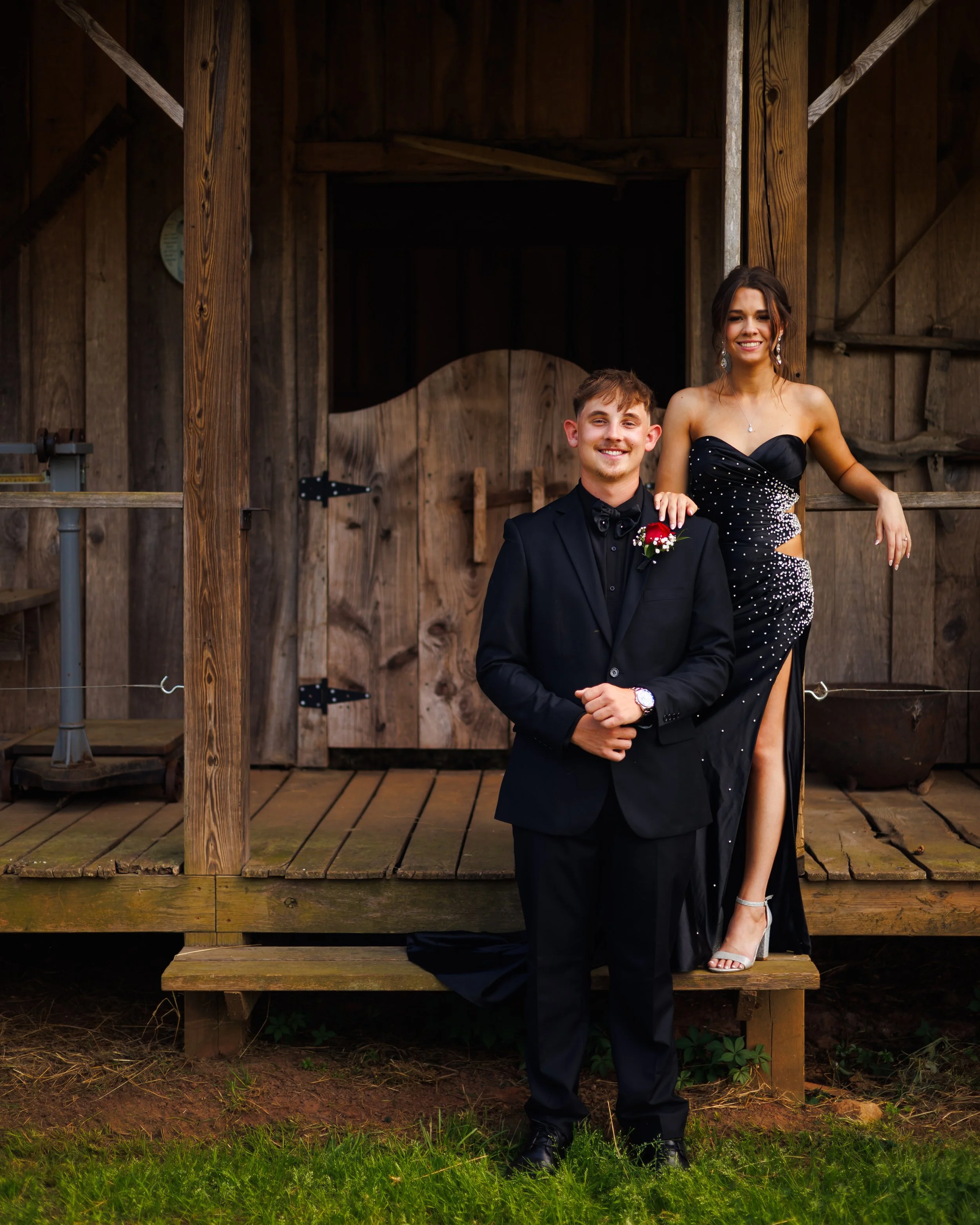 A young man in a black tuxedo and a young woman in a black dress with silver embellishments are posing together on a wooden porch. The woman is standing behind the man with her hand on his shoulder, both smiling at the camera. The setting appears to 