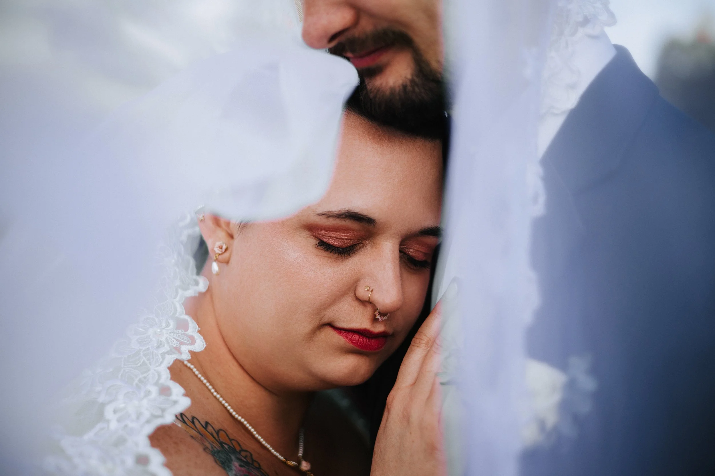 A close-up of a bride with closed eyes, wearing makeup, gold earrings, a nose ring, and a white lace wedding dress, touching her face with her finger.