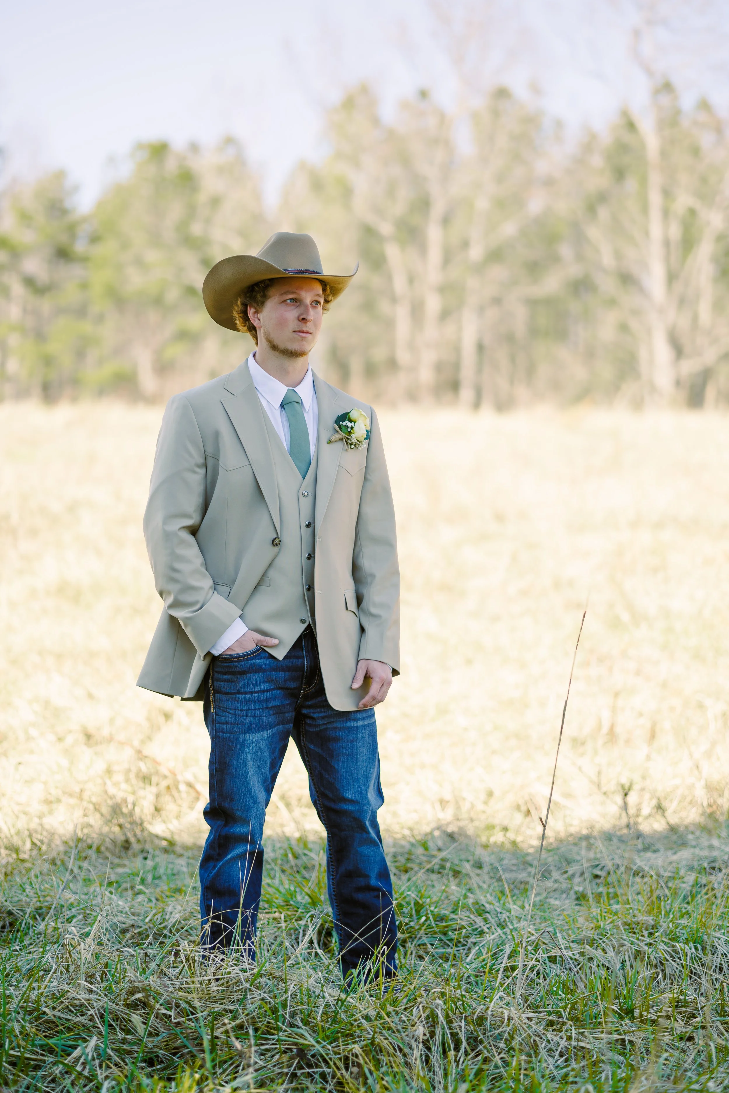 A young man wearing a beige blazer, white shirt, teal tie, blue jeans, and a cowboy hat standing in a grassy field with trees in the background.