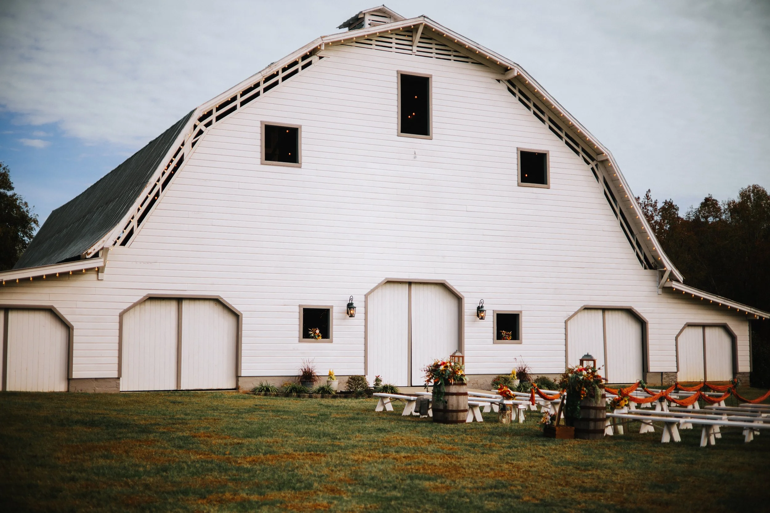 A white barn decorated for a special event with floral arrangements, lanterns, and string lights, set on a grassy lawn with trees in the background.