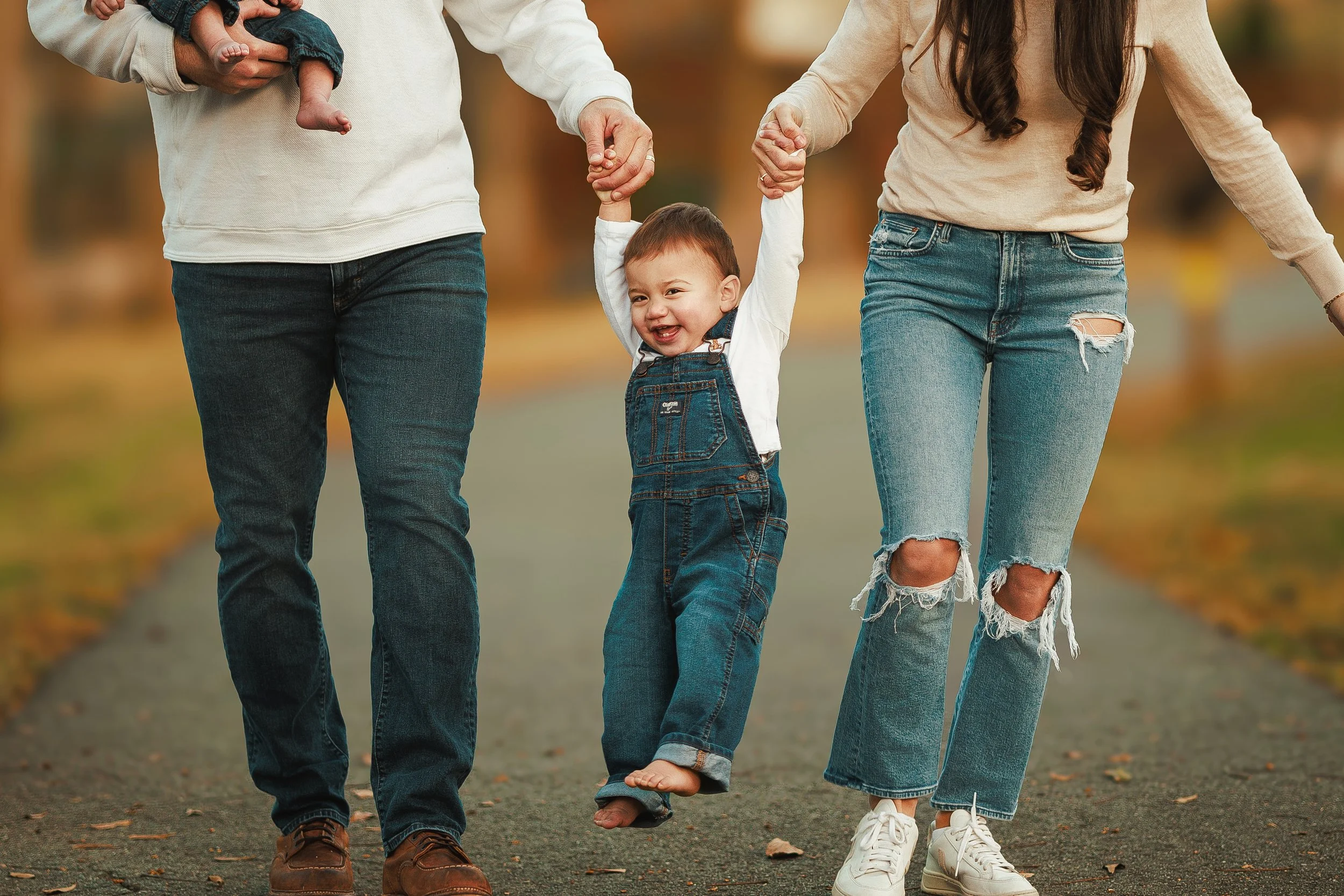 A family walking outdoors with a toddler. The child is holding hands with both parents, who are dressed casually. The scene is set in autumn with fall leaves scattered on the ground.