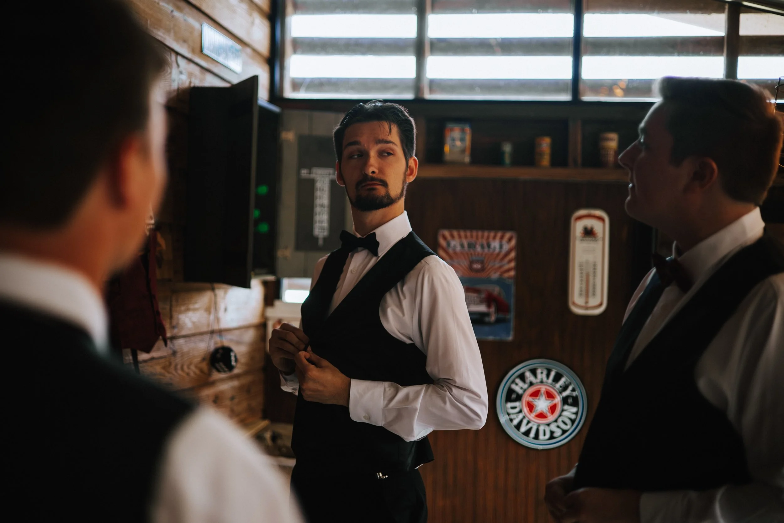 A man dressed as a waiter in a tuxedo with a bow tie, looking to his left while adjusting his shirt cuffs, in a setting with wooden walls and a Harley Davidson sign on the wall. Two other men wearing tuxedos are partially visible in the foreground.