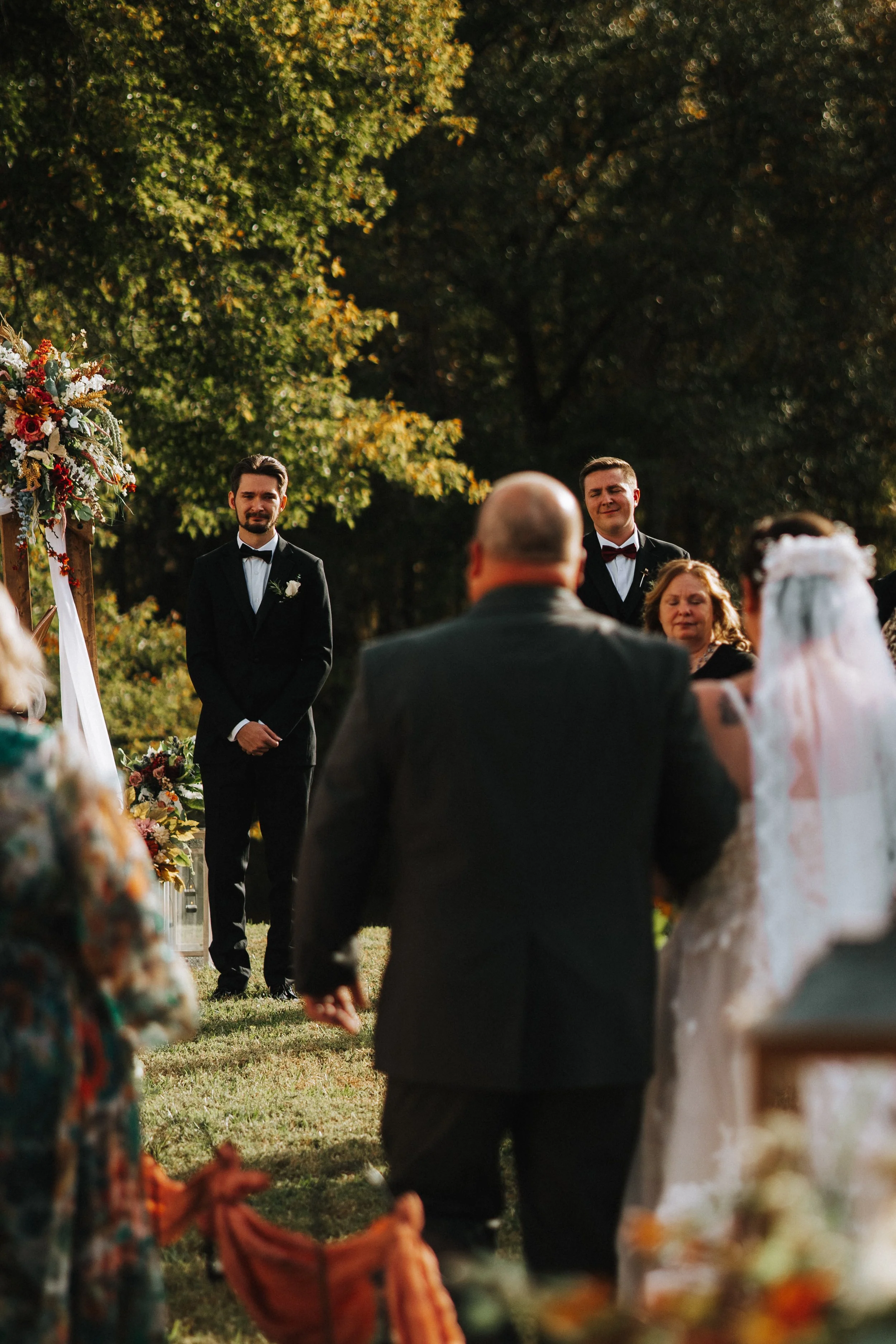 A couple of men dressed in formal suits during an outdoor wedding ceremony, with guests watching and a floral arch on the left side.