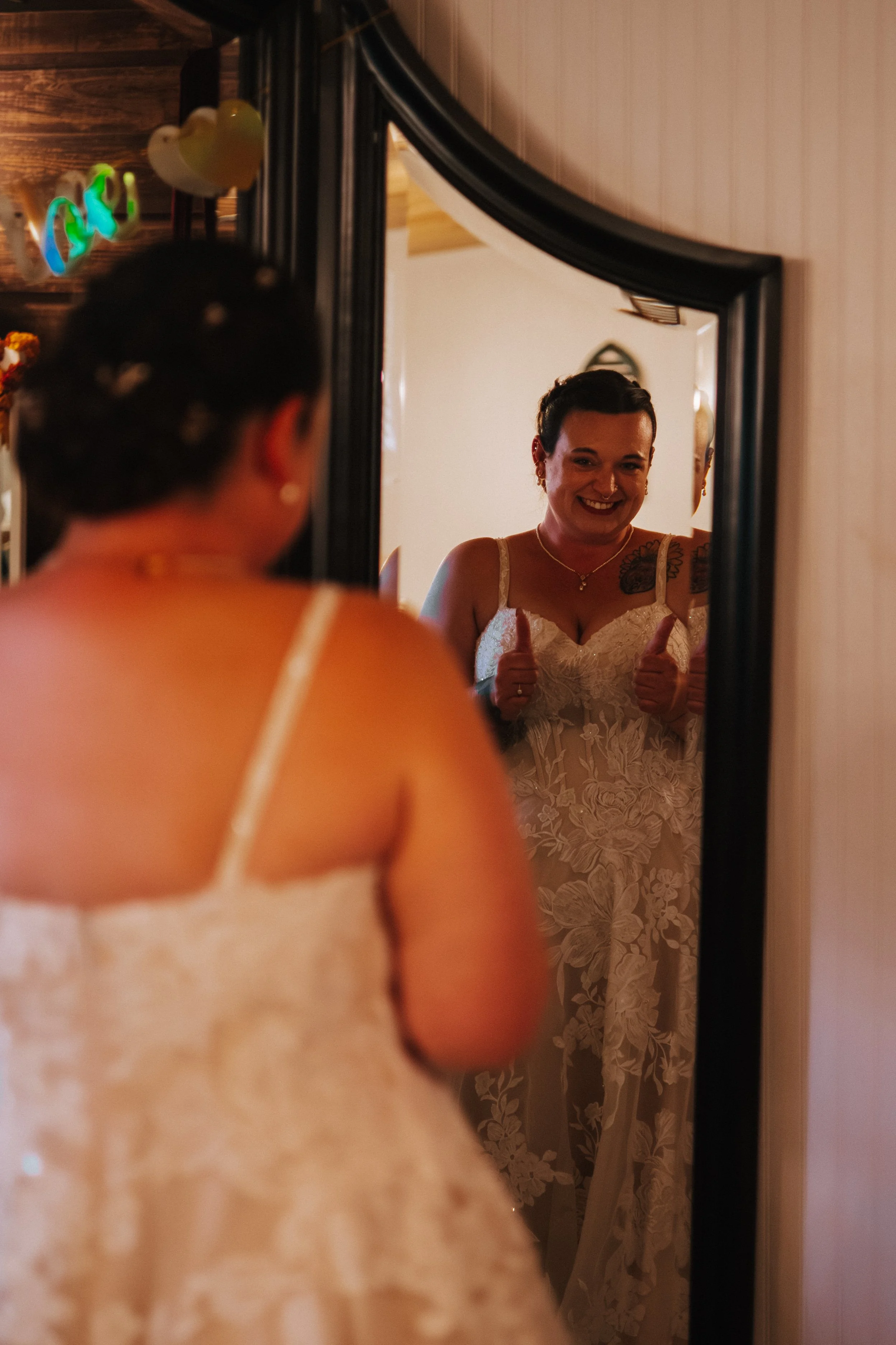 A woman with short dark hair and earrings looking into a mirror, giving thumbs up, wearing a white lace dress with floral patterns.