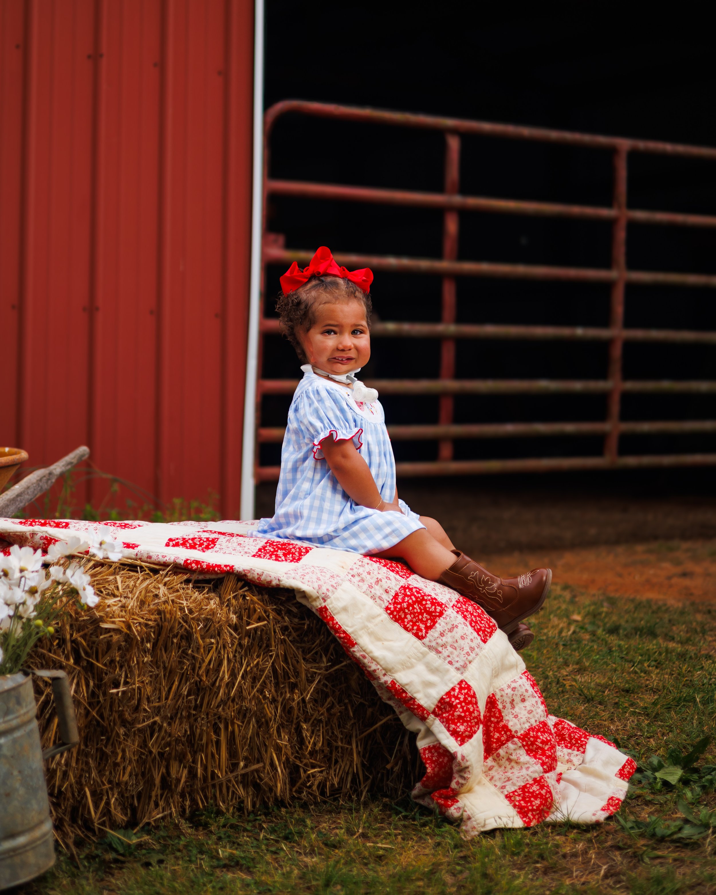 Young girl with curly hair wearing a red bow, blue plaid dress, and brown cowboy boots, sitting on a quilted blanket draped over hay bales, with a red barn and rusted farm equipment in the background.