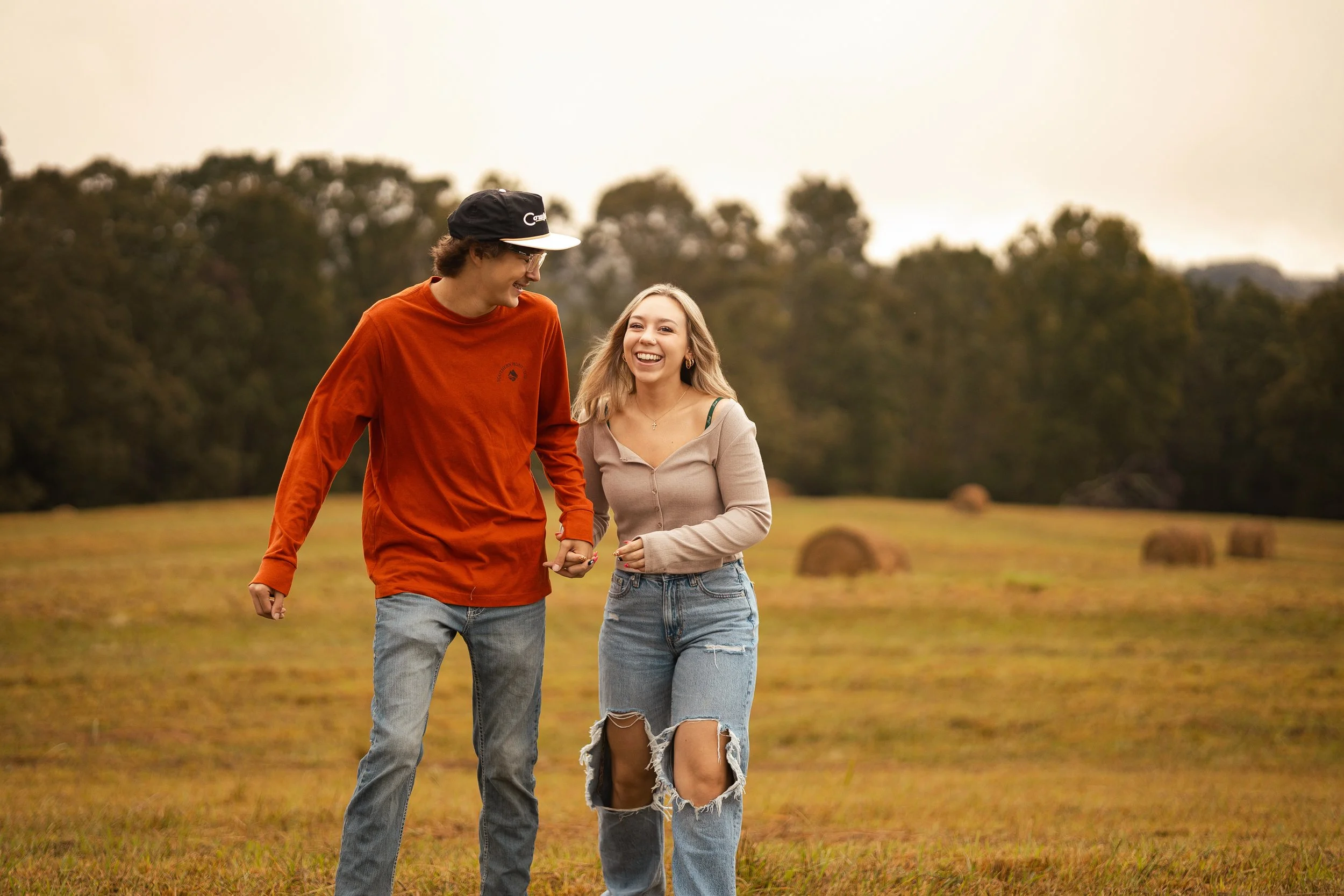 A young man and woman walking hand in hand outdoors in a field with hay bales, smiling and enjoying each other's company on a cloudy day.