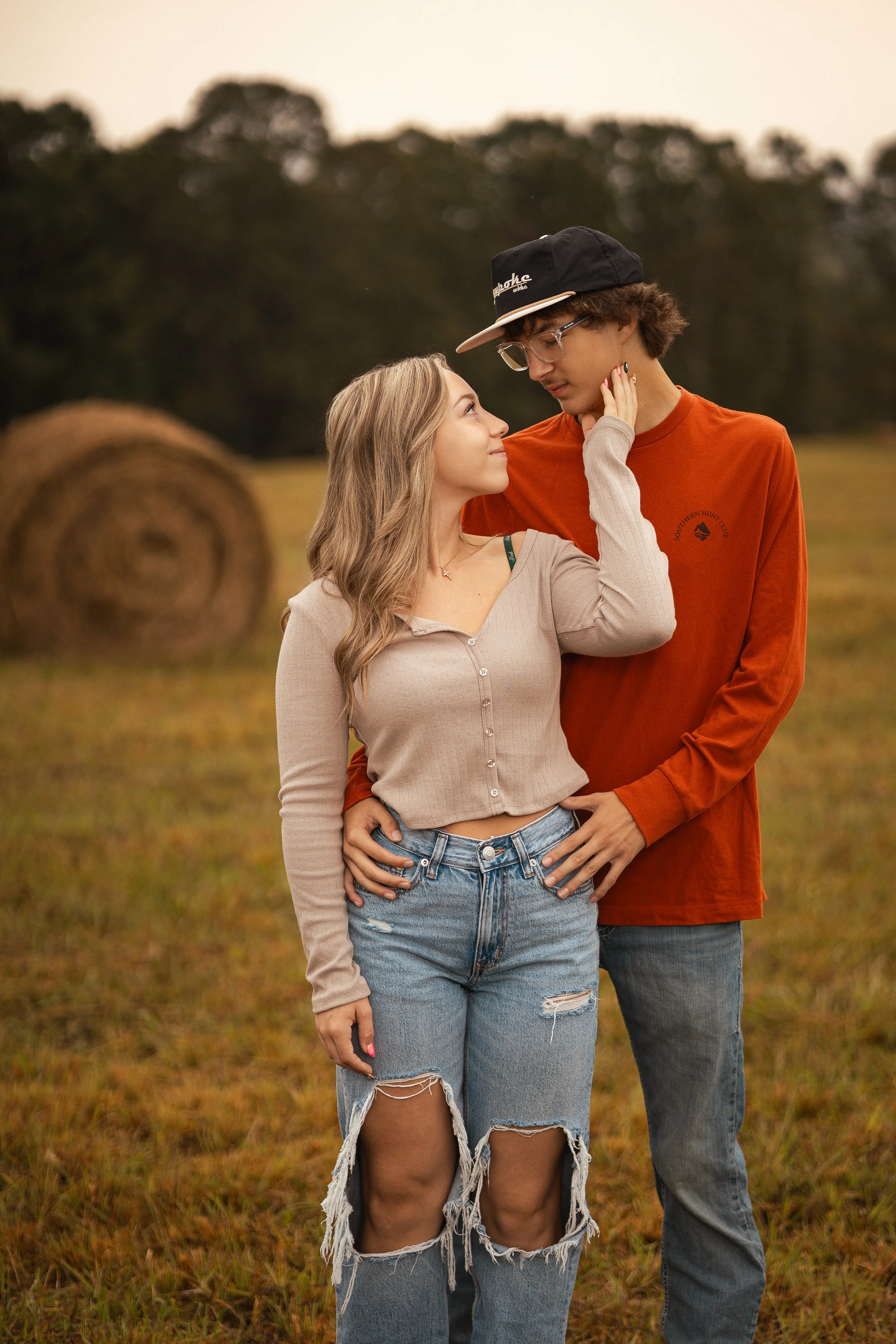 A young couple stands close together in a field, gazing into each other's eyes, with hay bales and trees in the background during daytime.
