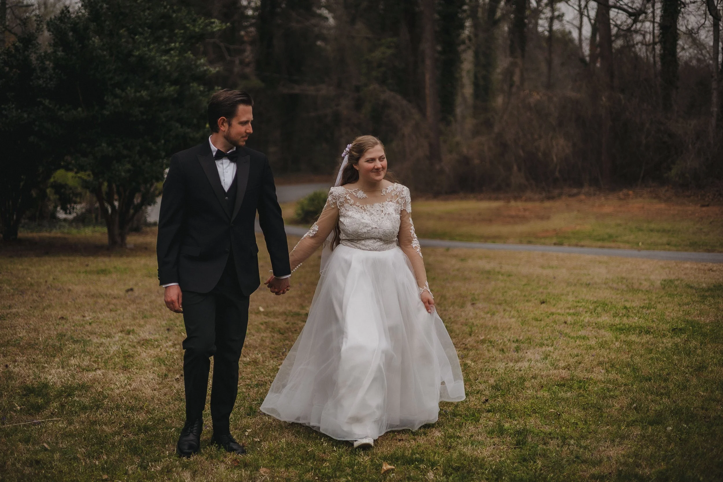 A bride and groom walking hand in hand on a grassy area outdoors during a wedding. The groom is dressed in a black tuxedo with a bow tie, and the bride is wearing a white wedding dress with lace details and a veil. They are smiling and looking at eac