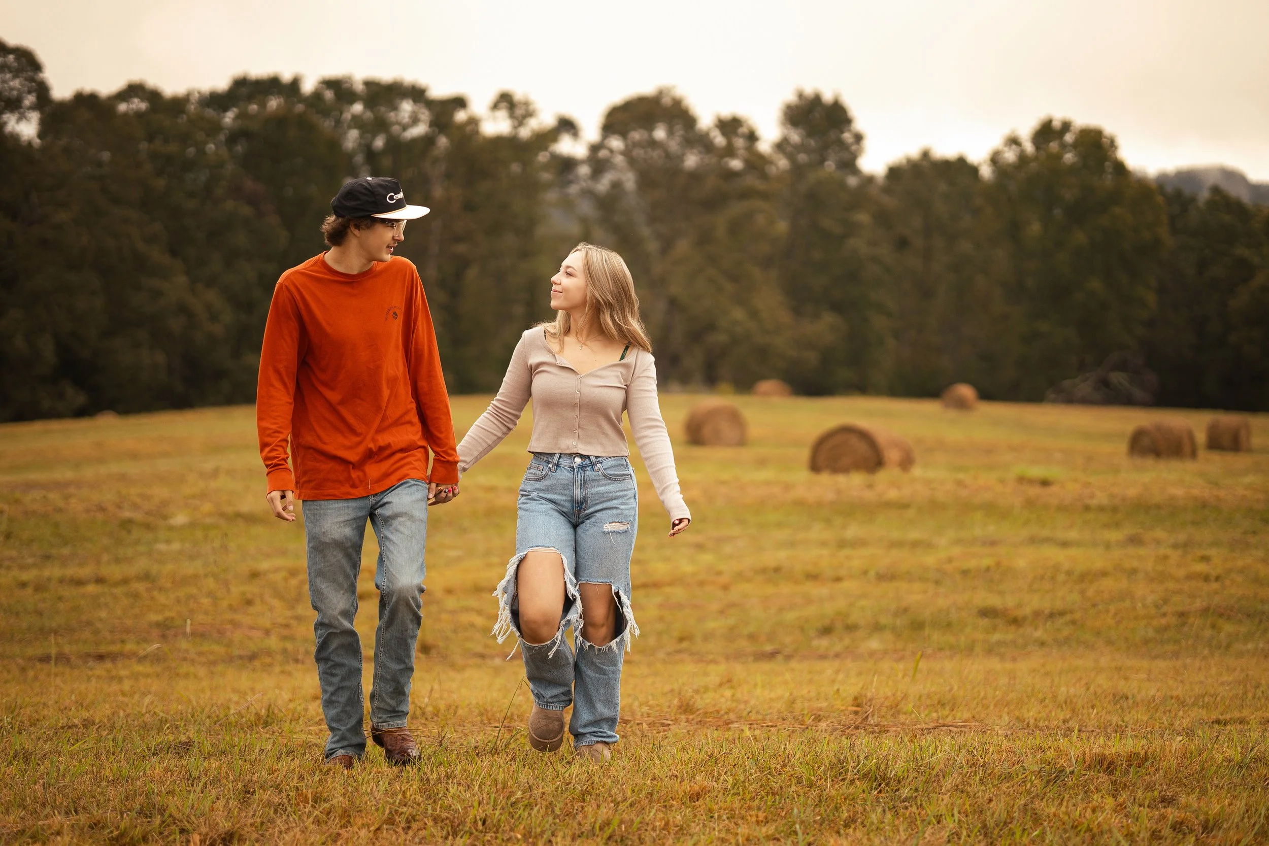 A young man and woman walk hand in hand in a grassy field with hay bales and a line of trees in the background, during what appears to be late afternoon or early evening.