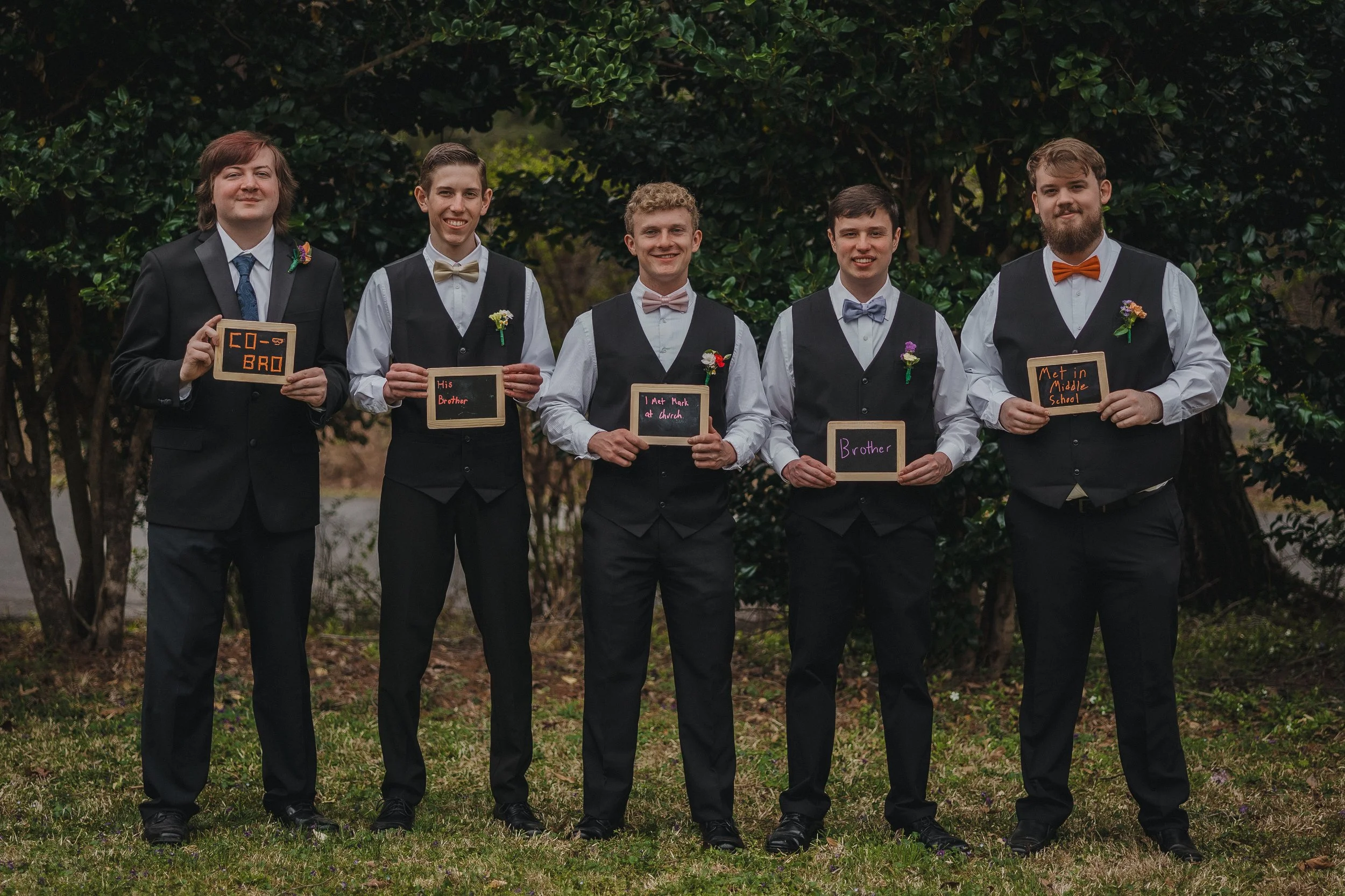 A group of five young men standing outdoors in front of bushes, dressed in formal attire with vests and bow ties, each holding a small sign with labeled personal details.