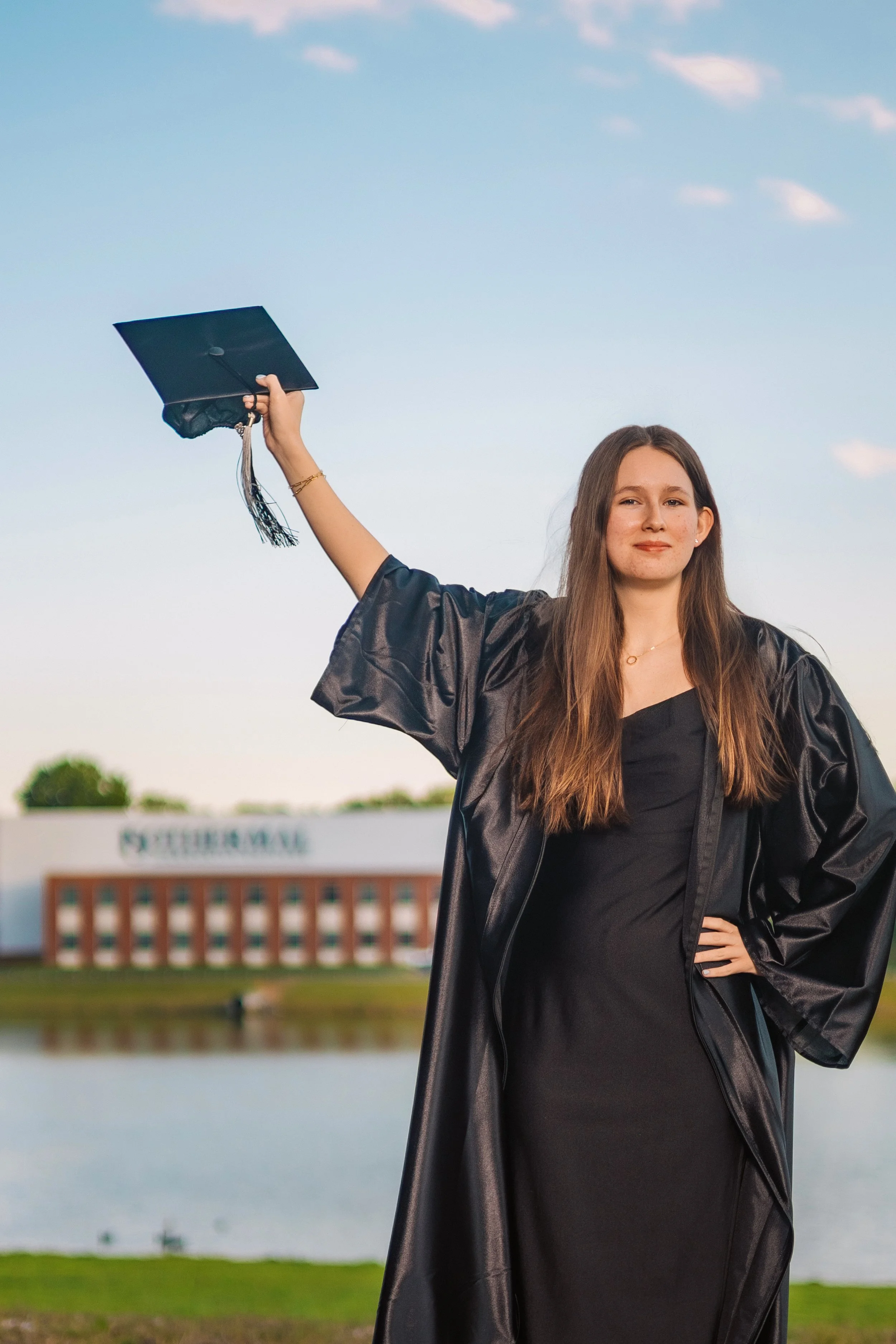 A young woman in graduation attire holding her cap in the air outdoors with a building and water behind her.