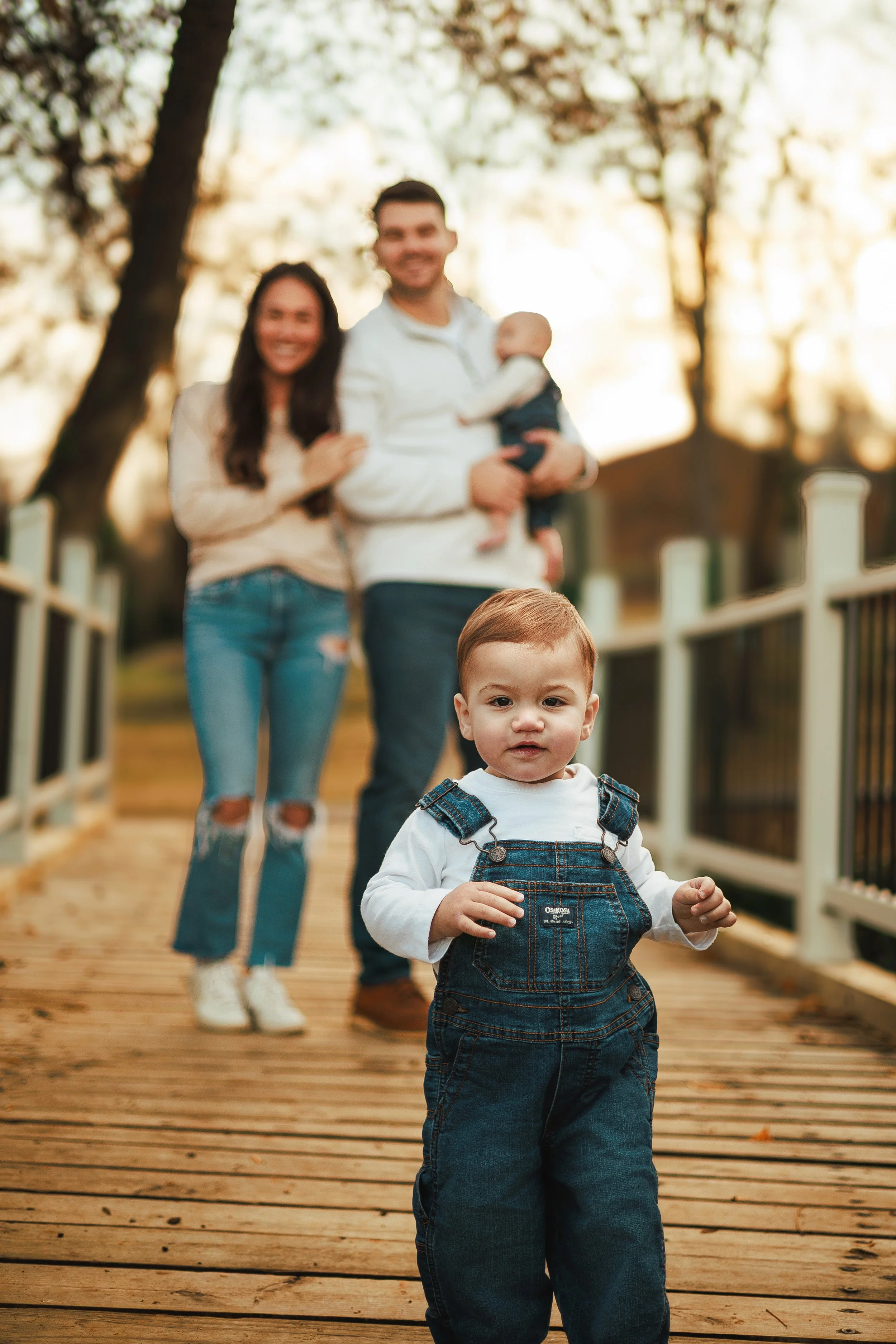 A young boy with short hair in denim overalls and a white long-sleeve shirt walking on a wooden bridge, with a family in the background, all outdoors surrounded by trees during sunset or golden hour.
