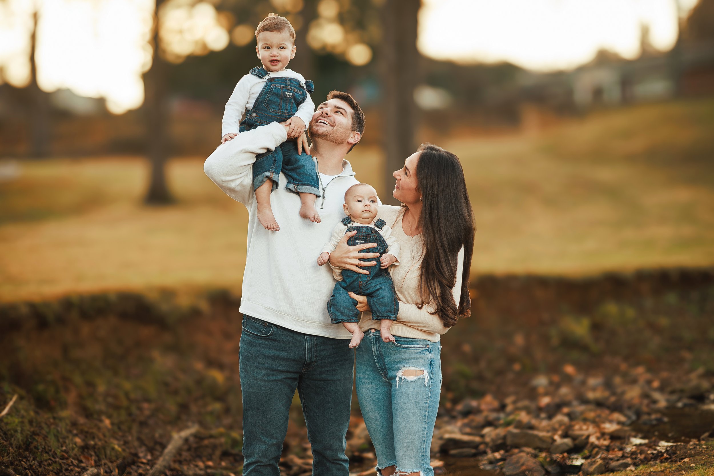 A man holding a young boy on his shoulder and a woman holding a baby, walking outdoors in a park during fall with trees in the background.