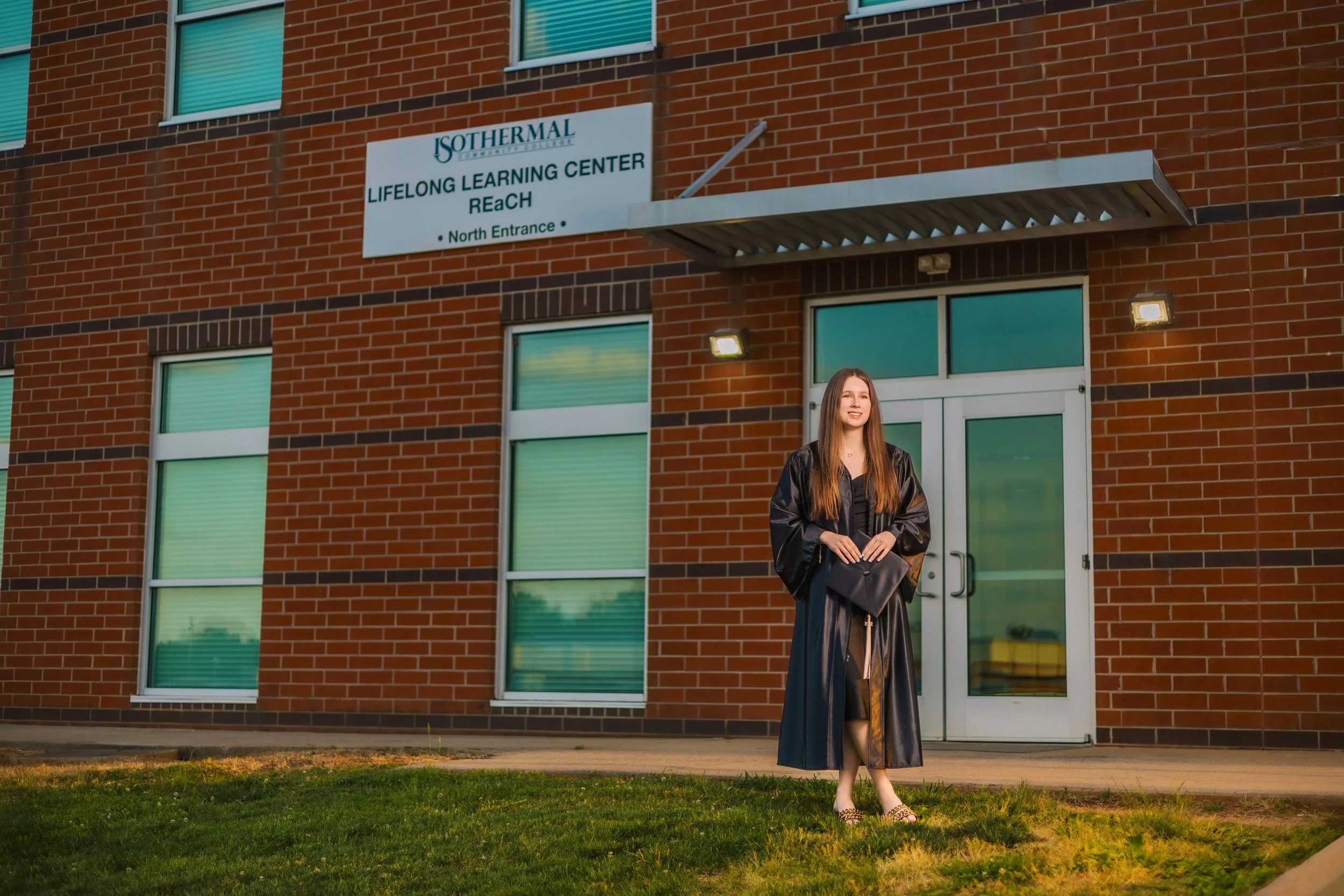 A young woman in graduation gown standing recording device in front of a brick building with a sign that reads 'LIFELONG LEARNING CENTER ReACH North Entrance'.