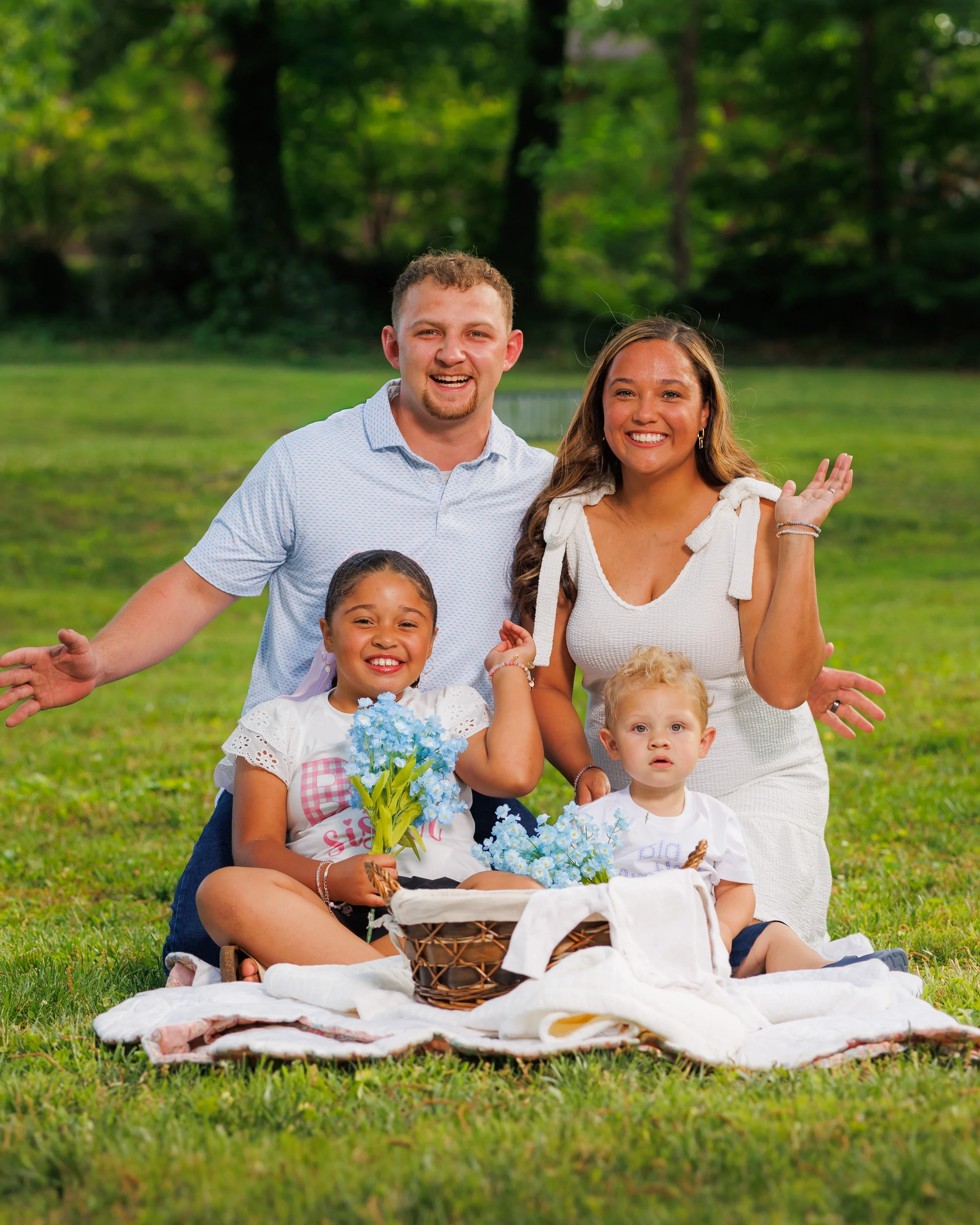 Family of five having a picnic on a blanket in a park with green grass and trees in the background, smiling and enjoying each other's company.