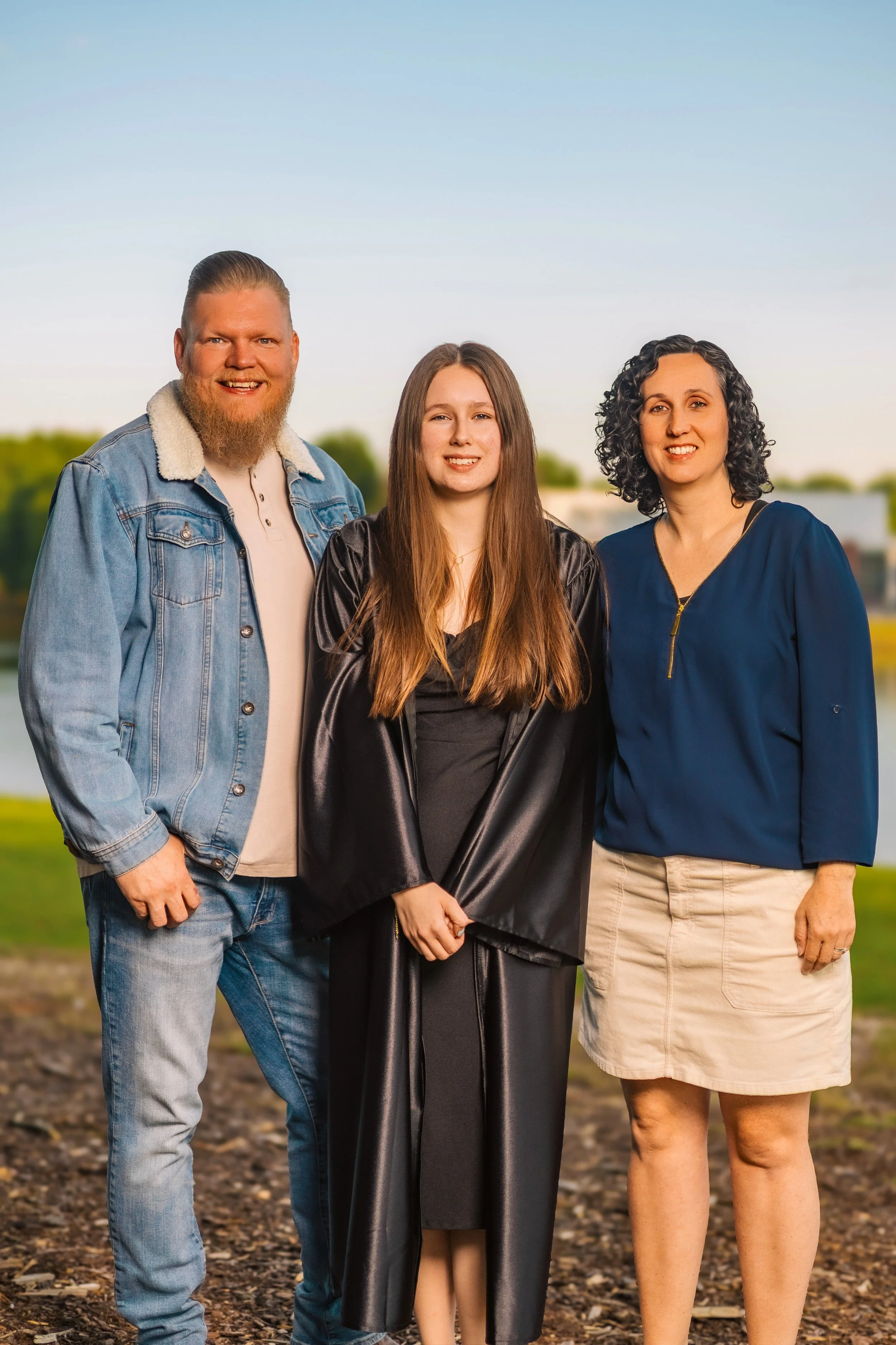 A group of three people standing outdoors on a dirt trail with greenery and a blue sky in the background, smiling at the camera. The group consists of a man with a beard in a denim jacket, a woman with long hair in a black graduation gown, and a woma