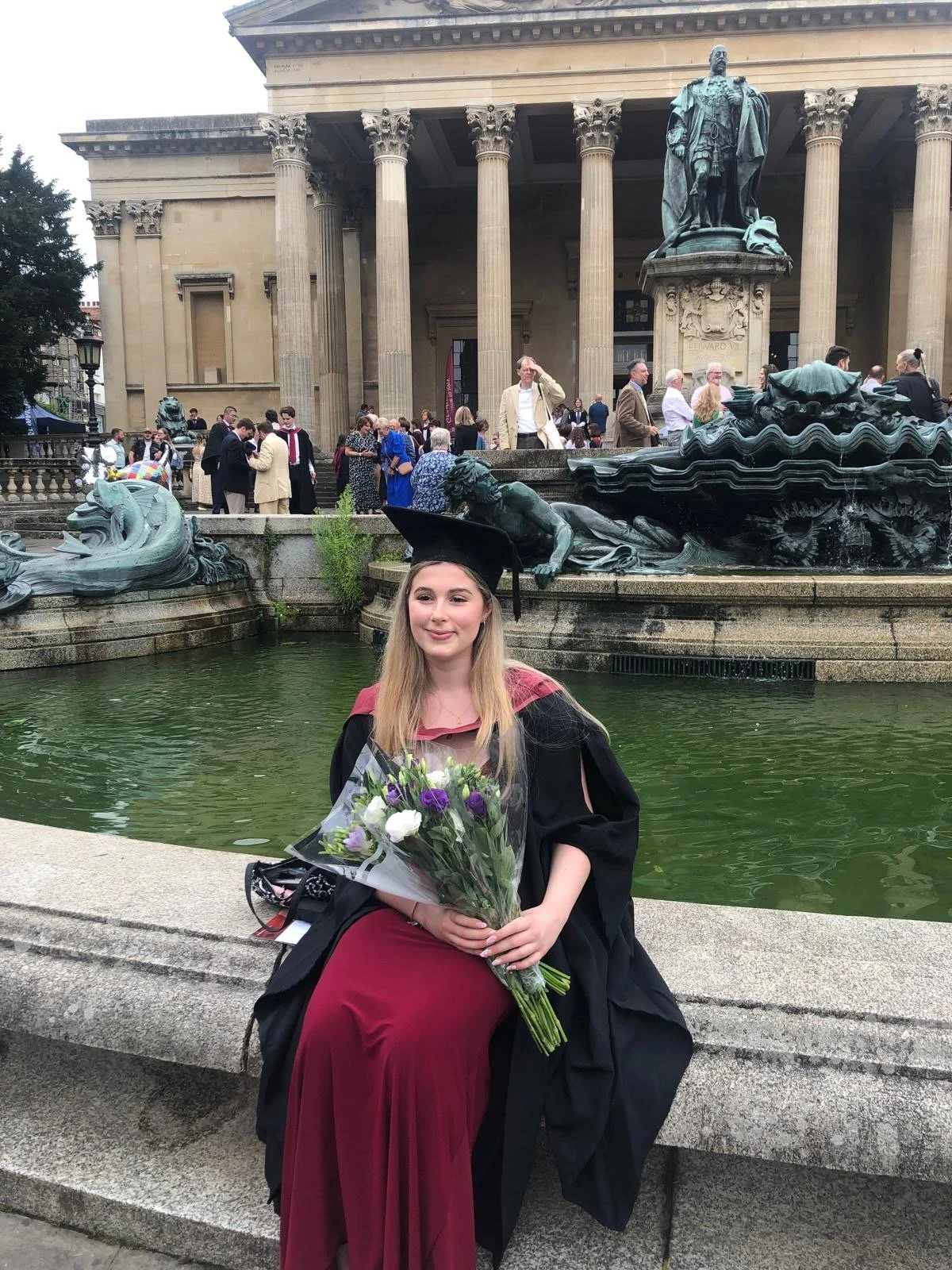 A young woman in a graduation gown and cap sitting on a stone bench, holding a bouquet of flowers in front of a fountain with historical sculptures, and a large neoclassical building with columns and a statue in the background.