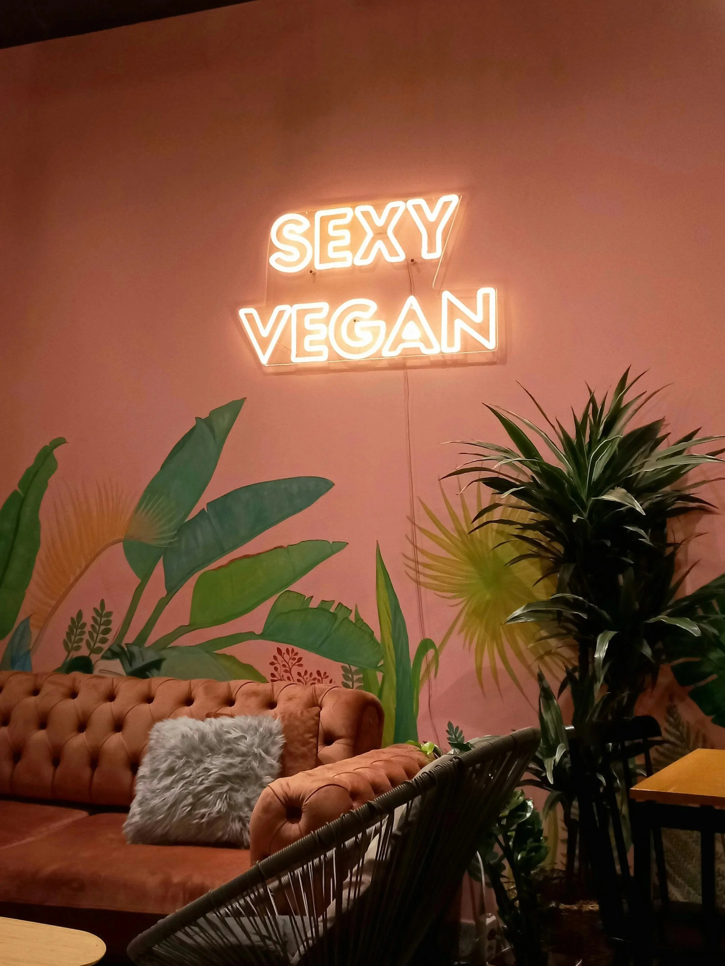 A pink wall with a neon sign that says 'SEXY VEGAN.' In front are green tropical plants, an orange tufted sofa with a gray fluffy pillow, and part of a black rattan chair.