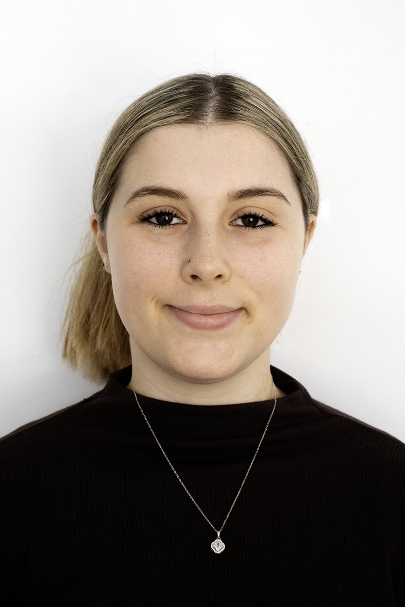 A young woman with light brown hair in a ponytail, wearing a black turtleneck sweater and a silver necklace with a pendant, standing against a white background.