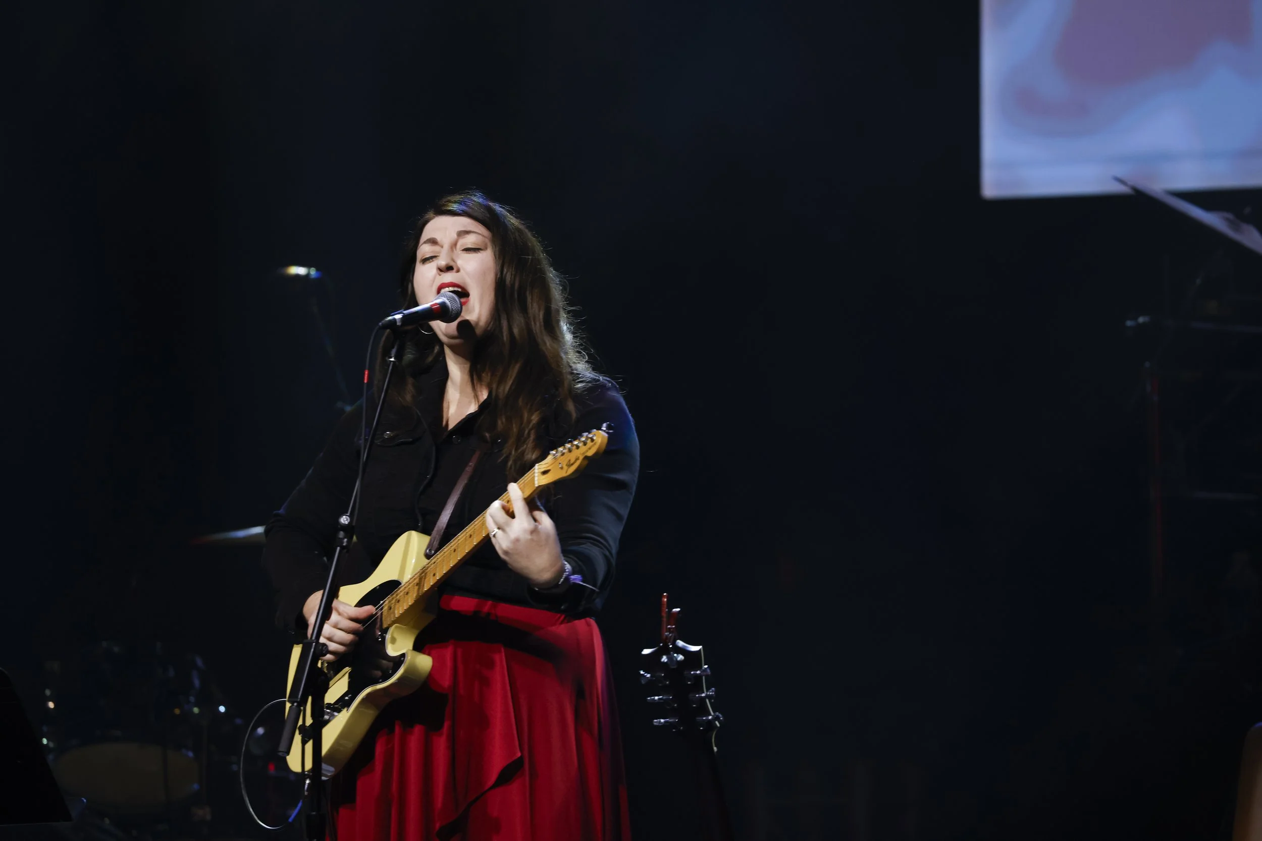 A female musician singing into a microphone while playing an electric guitar on stage during a performance.