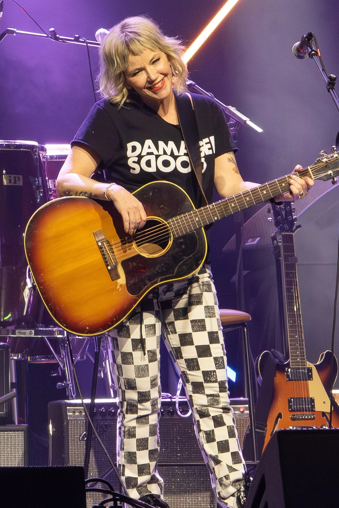 A woman smiling and playing an acoustic guitar on stage with purple lighting, wearing a black T-shirt with text and checkered pants, with additional musical equipment visible in the background.