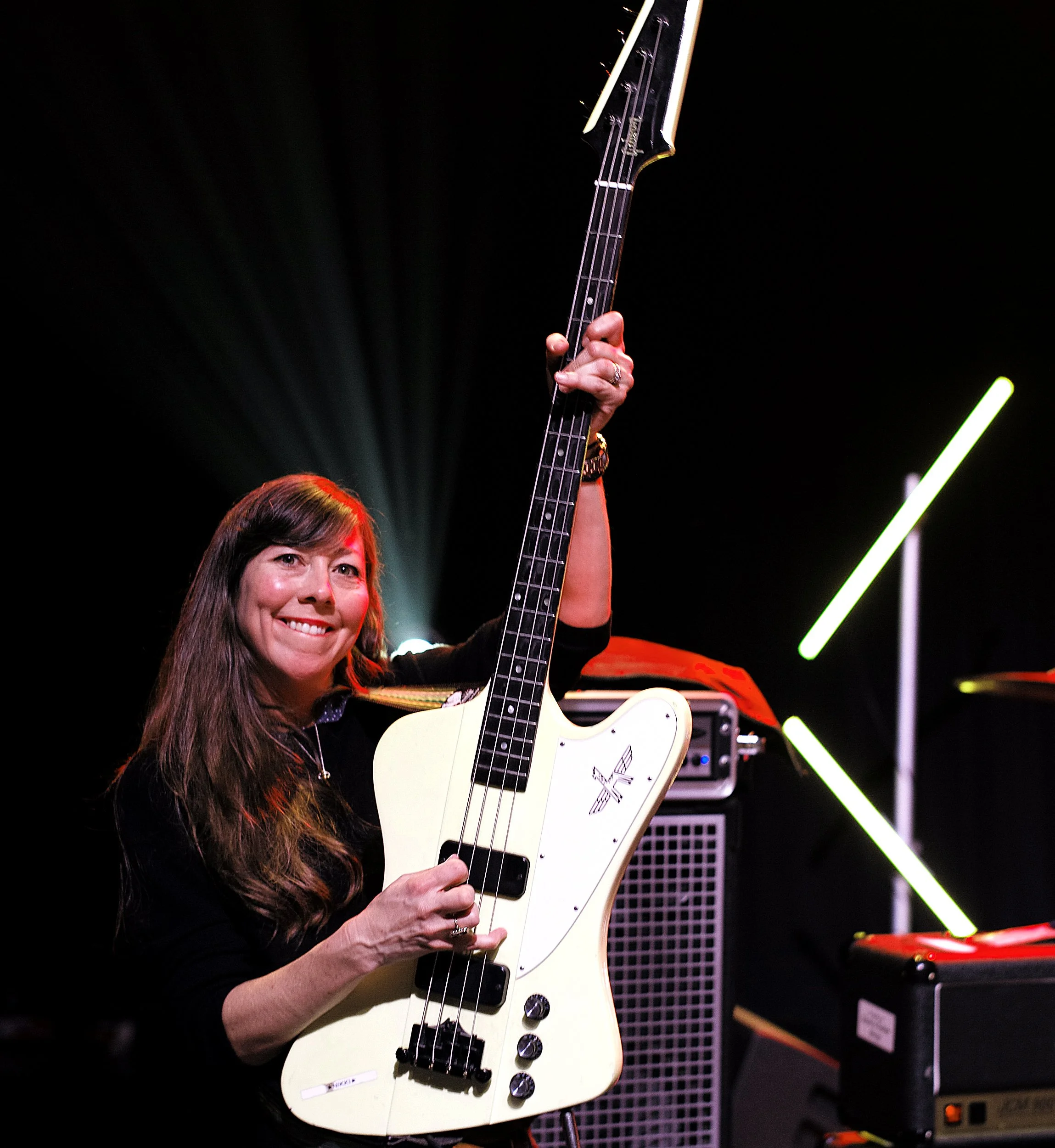 A woman with long brown hair holding a white electric bass guitar on a stage with black background and neon light decor.