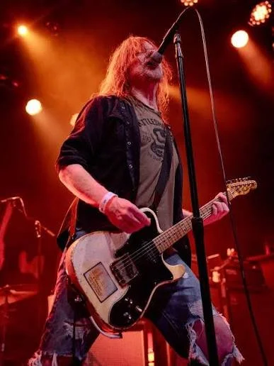 A musician with long hair performing on stage, singing into a microphone and playing an electric guitar, illuminated by colorful stage lighting.