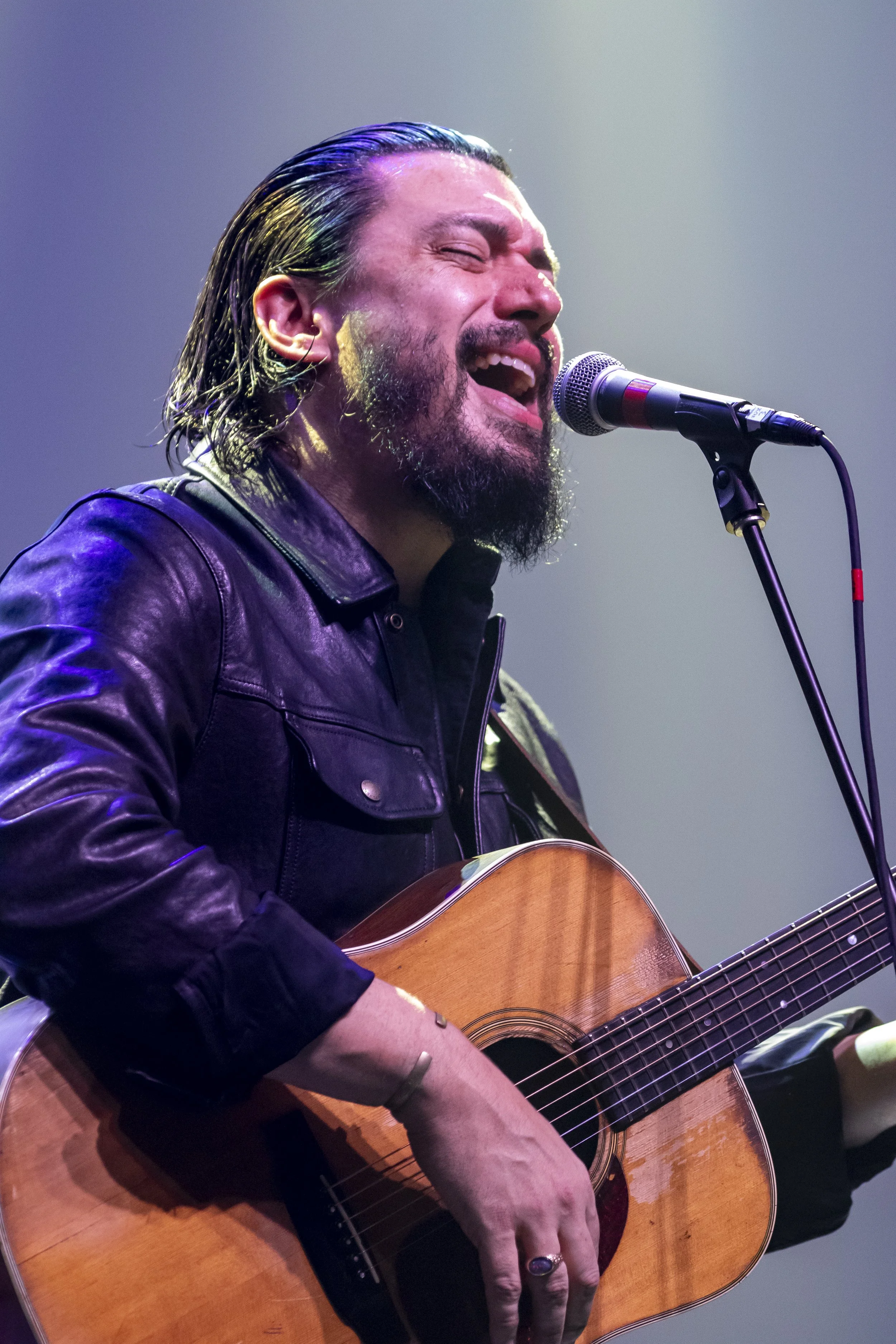 A man with long, dark hair and a beard passionately singing into a microphone while playing an acoustic guitar. He is wearing a black leather jacket and a ring on his finger. The background is blurred with colorful stage lighting.