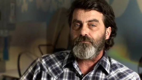 A middle-aged man with dark hair and a beard, wearing a plaid shirt, sitting indoors.