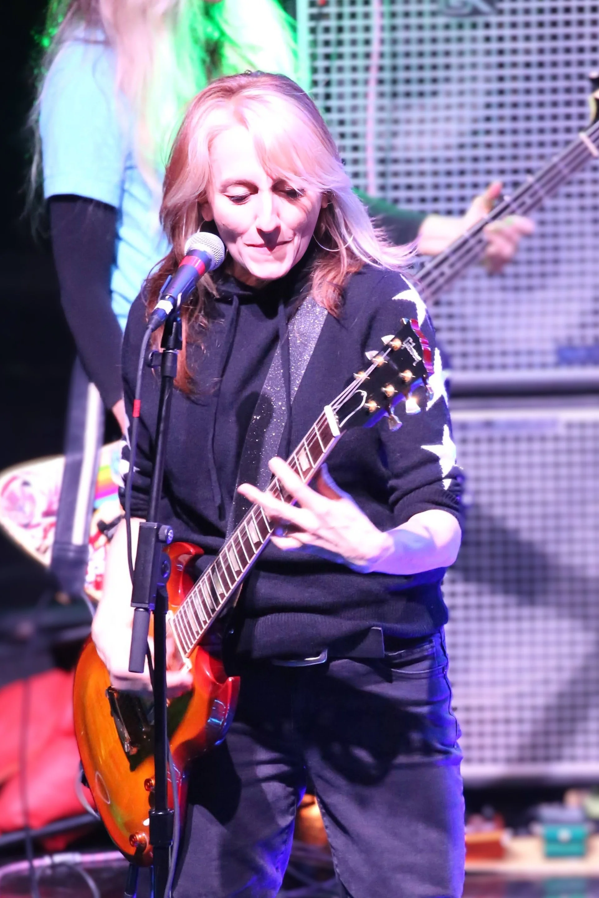 A woman with red hair playing an electric guitar and singing into a microphone on stage, with an amplifier in the background.