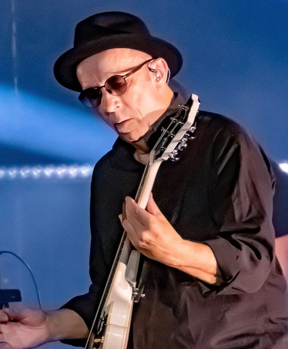 A man wearing a black hat and glasses playing an electric guitar on stage.