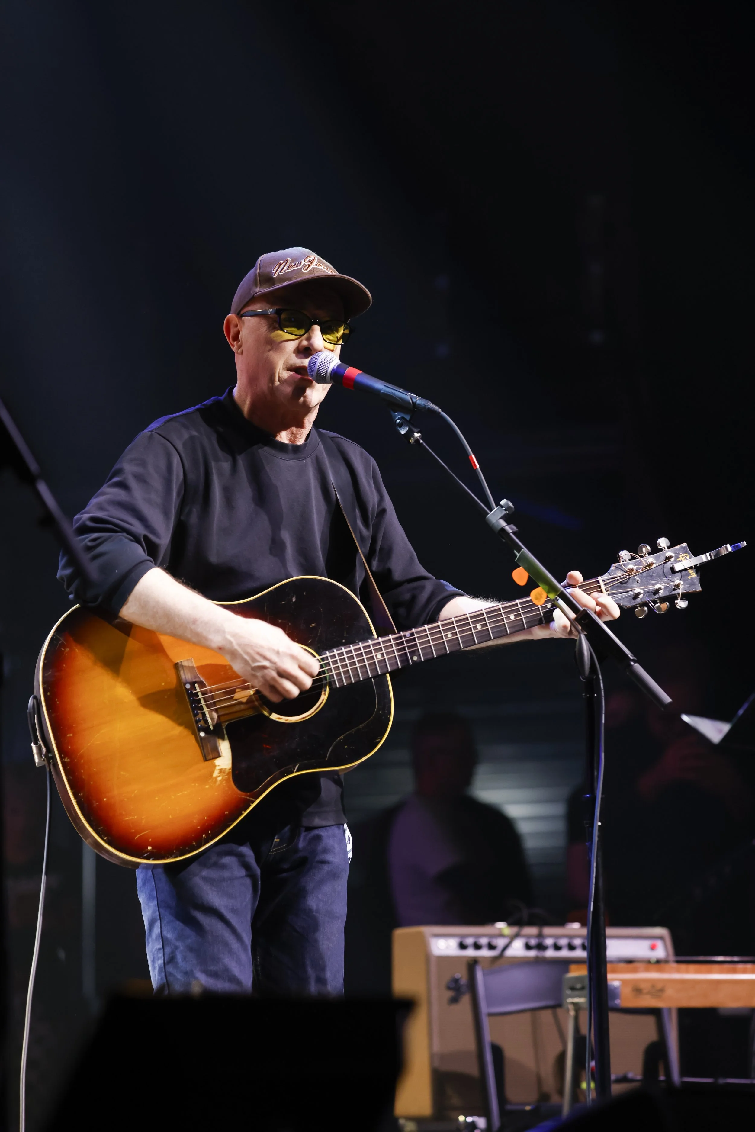 A man playing an acoustic guitar and singing into a microphone on stage, wearing a cap, glasses, and a black shirt.