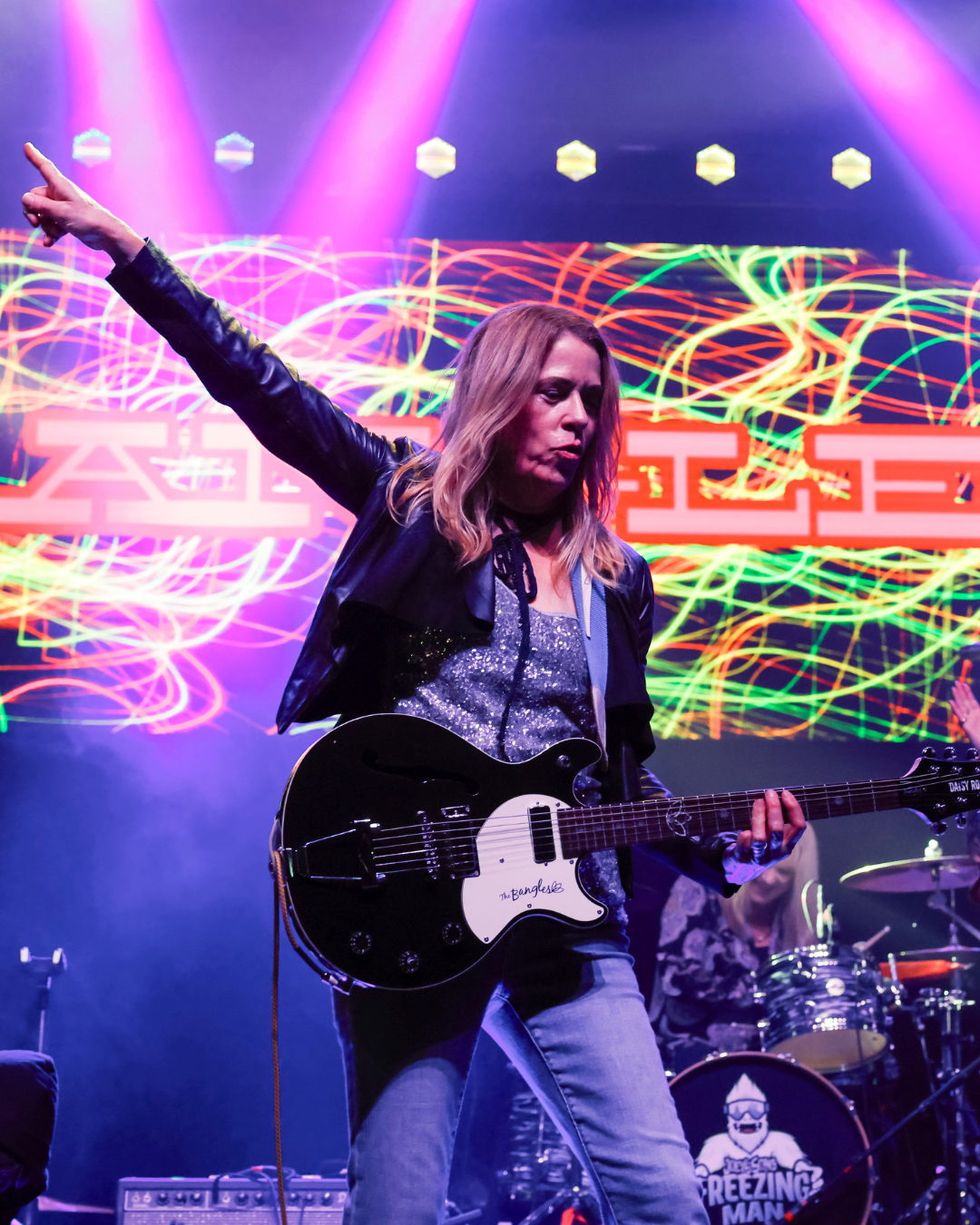 Musician playing an electric guitar on stage with colorful laser lights and a background screen displaying red and green graphics.
