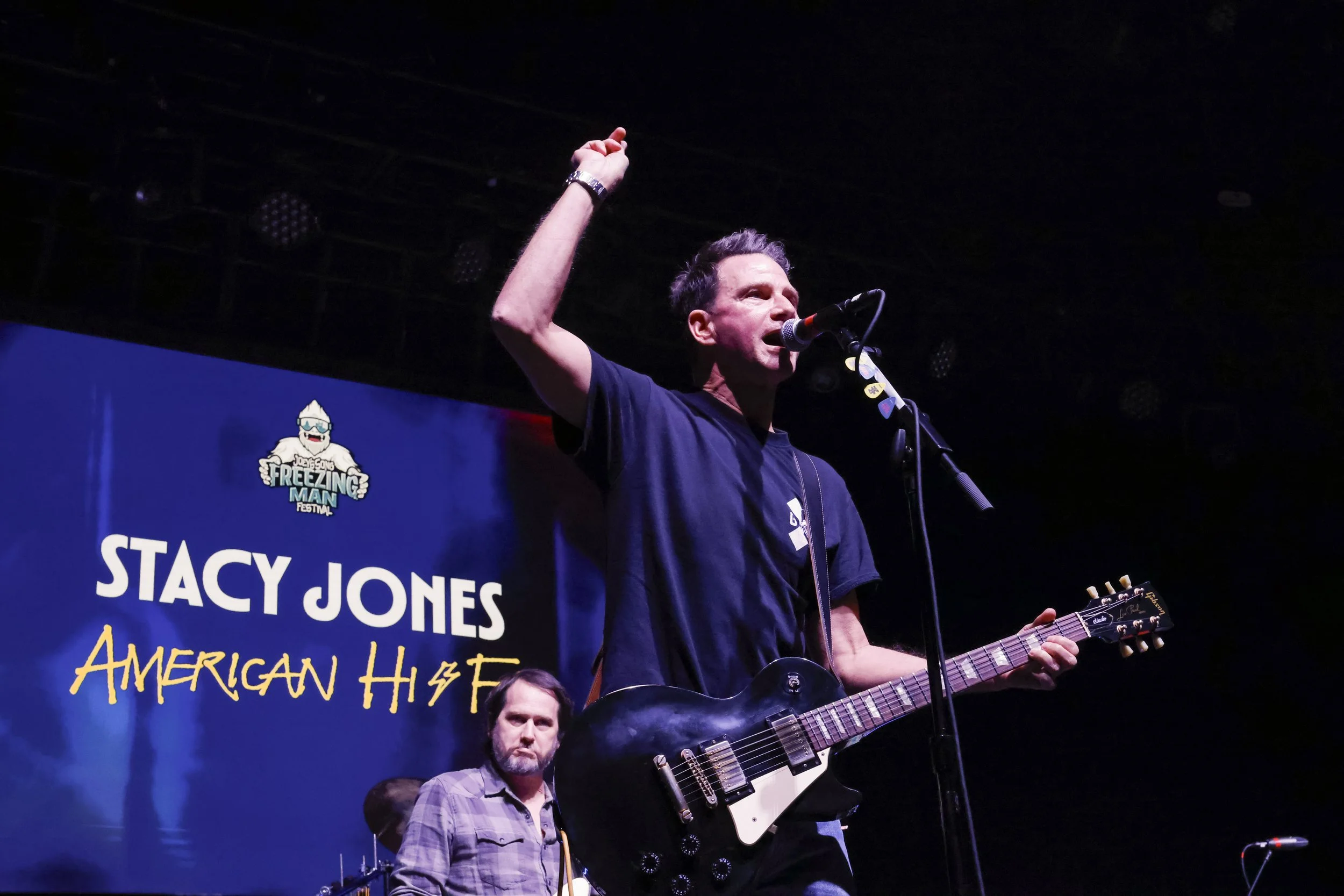 Man playing electric guitar and singing at a concert on stage at the American Hustle Music Festival, with a blue banner in the background featuring the names Stacy Jones and the event logo.