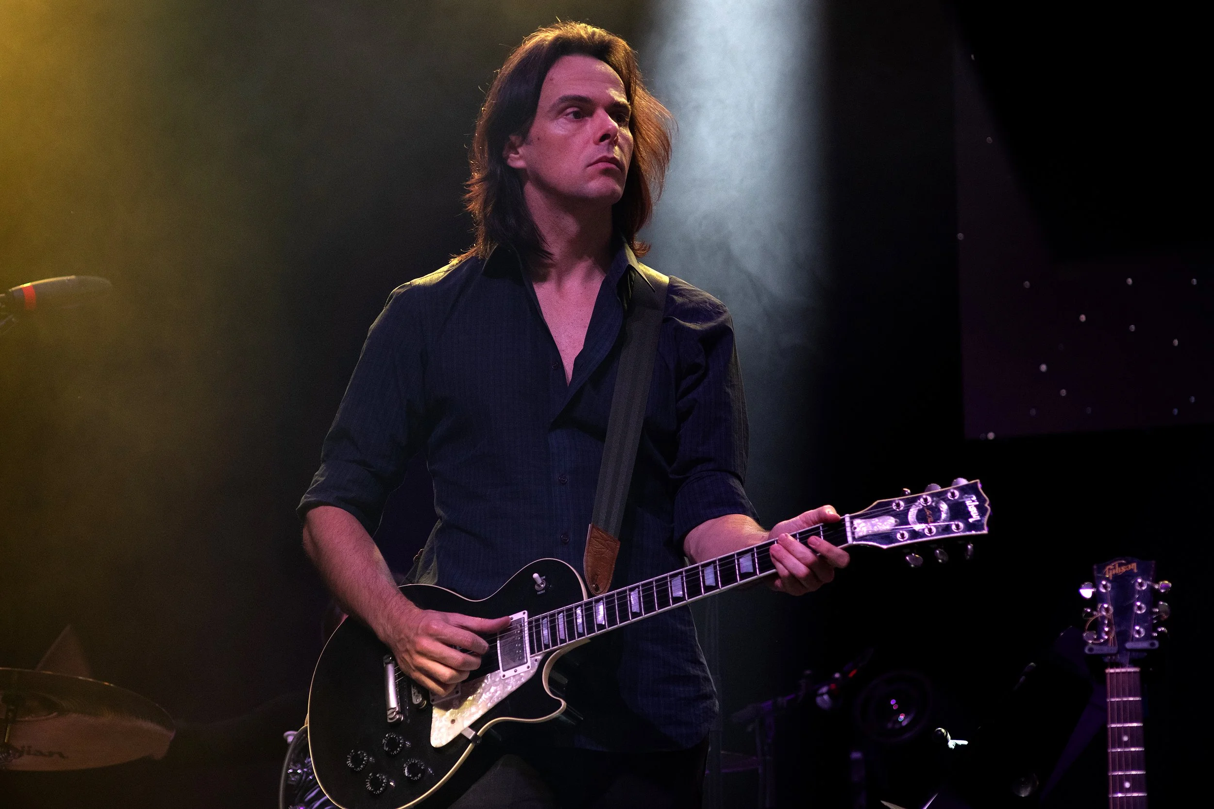 A man with long dark hair playing an electric guitar on stage, wearing a dark shirt, under dim stage lighting.