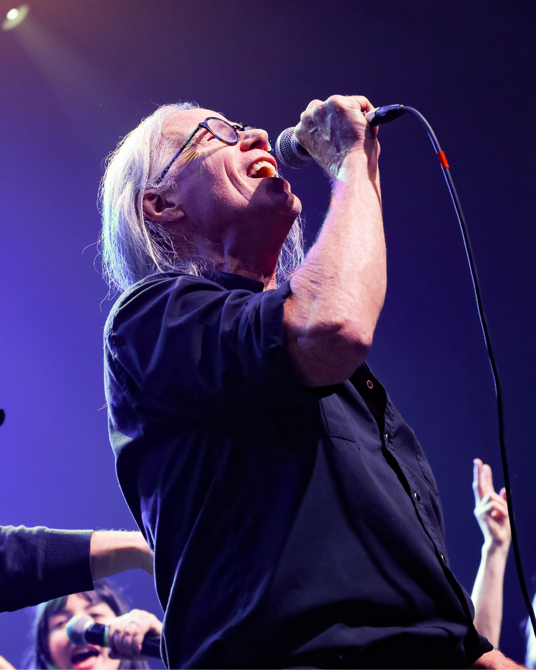 An elderly man with glasses and long gray hair singing passionately into a microphone on stage with purple and blue lighting.