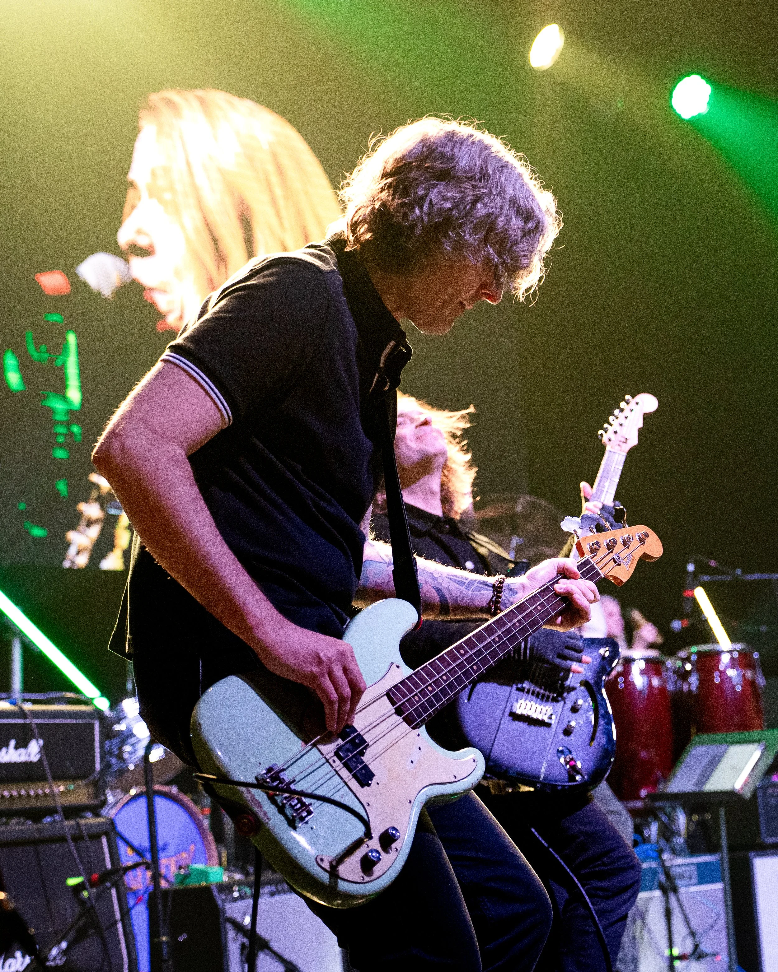 Guitarists performing on stage with colorful lighting, multiple guitars and musical equipment visible.