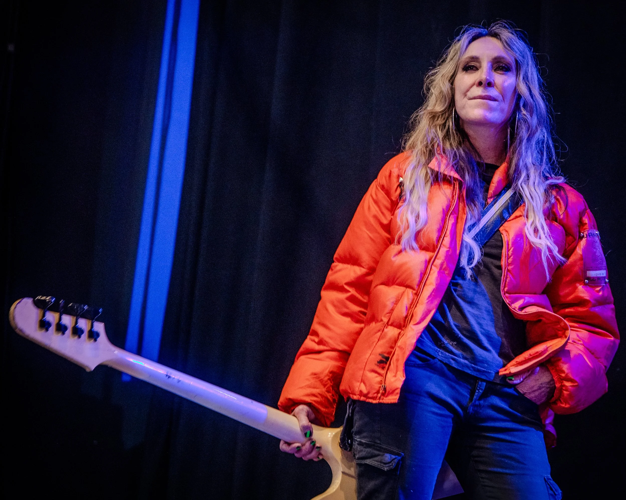 A woman with curly blonde hair and earrings wearing a bright orange jacket and black pants, standing against a dark background, holding a white electric guitar.