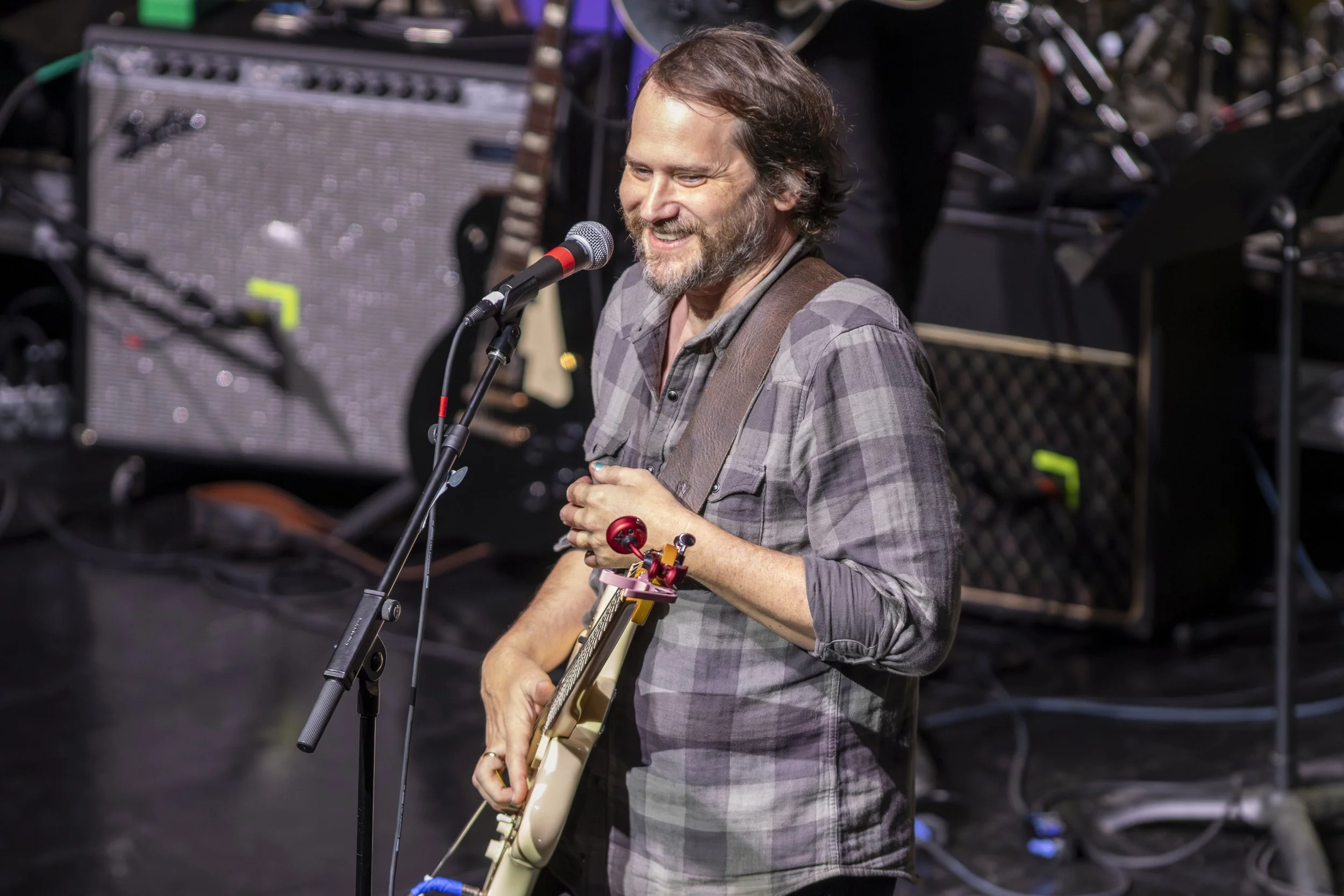 A man with a beard and long hair smiling while playing an electric guitar at a live music event. He is standing in front of music equipment and a microphone, wearing a plaid shirt and red wristband.