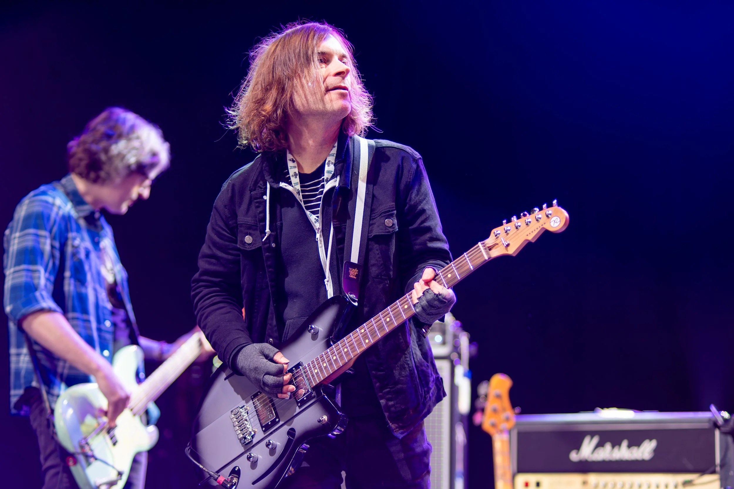 A musician with long hair playing an electric guitar on stage, with another guitarist visible in the background, in front of Marshall amplifier.