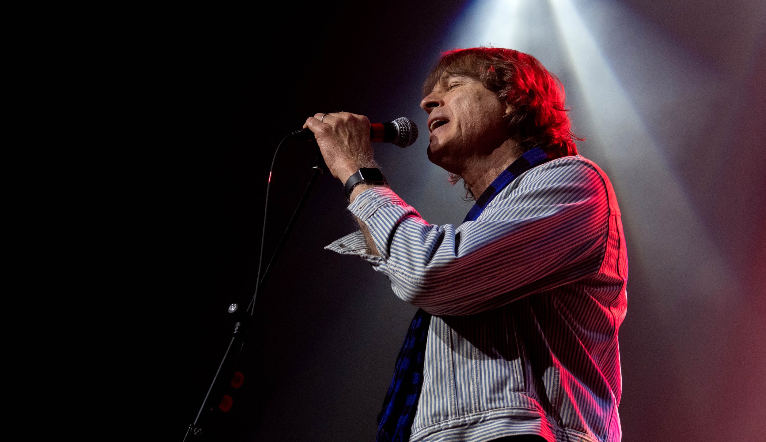 Adult male singer performing on stage with microphone, wearing a striped shirt, with stage lighting creating a dramatic effect.